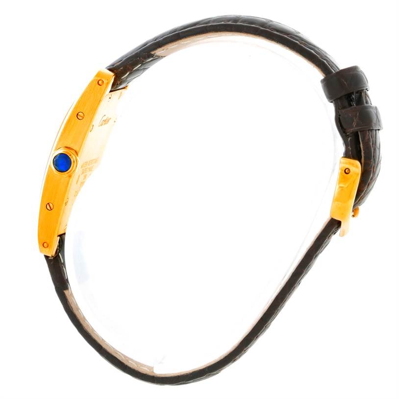 The image shows a side view of a Cartier Tank Americaine W2601956 Women's Yellow Gold Silver Dial W2601956 Women's Yellow Gold Silver Dial watch, highlighting its gold case, crown with a blue jewel, and leather strap.