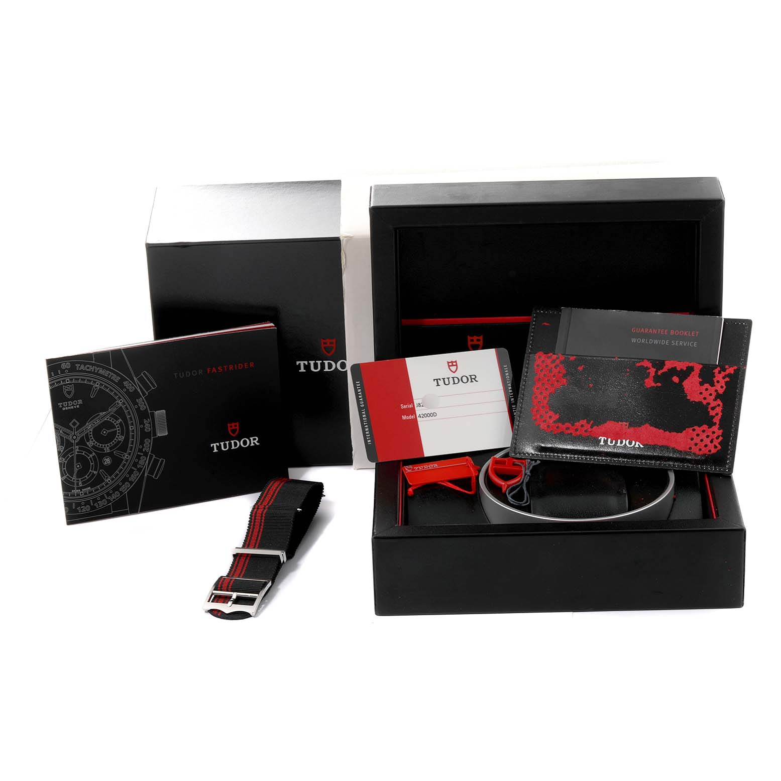 The image shows a Tudor Fastrider 42000D Men's Stainless Steel Red Dial 42000D Men's Stainless Steel Red Dial watch box set including a strap, warranty cards, manuals, and packaging from a high-angle view.