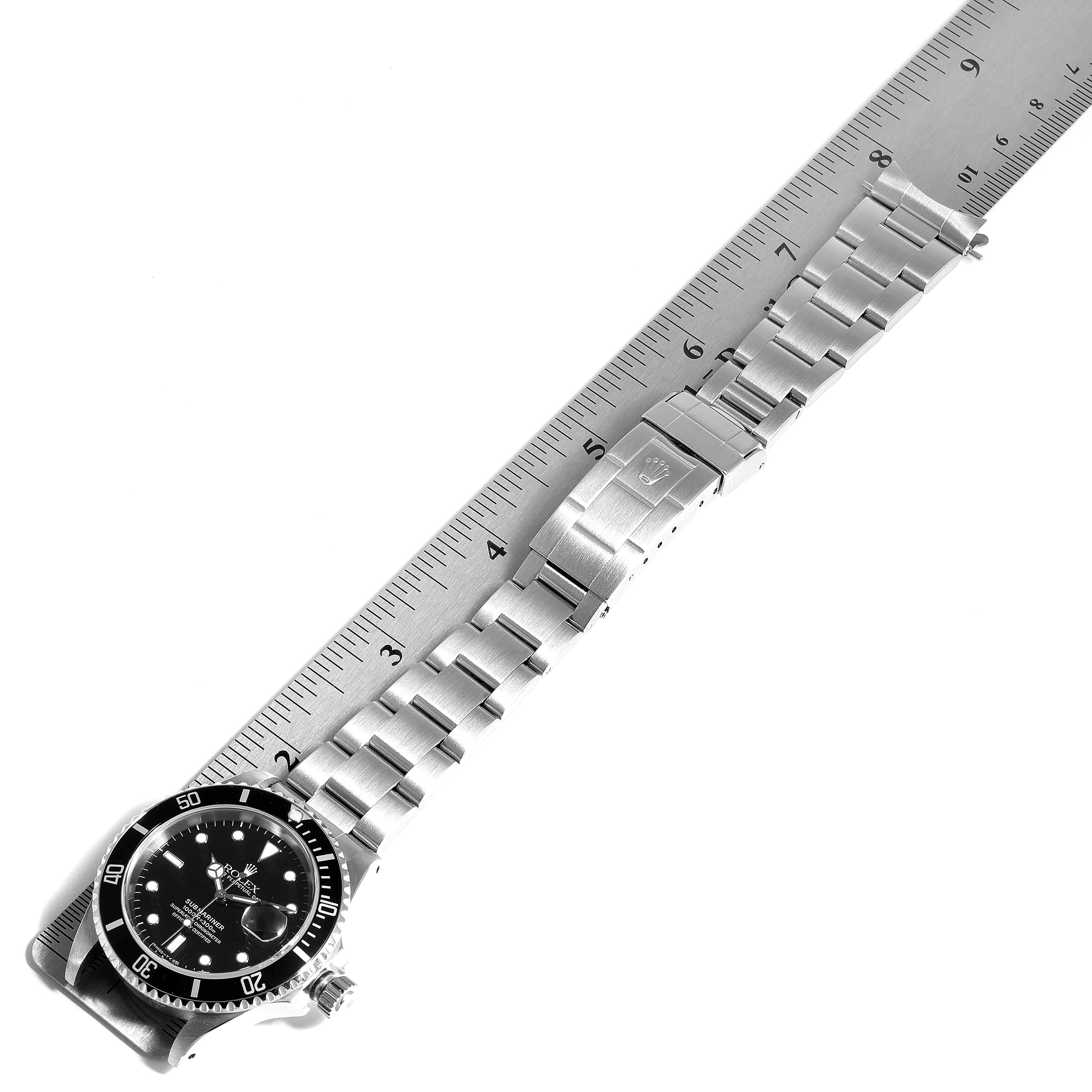 The image shows a Rolex Submariner 16610 Men
s Stainless Steel Black Dial 16610 Men
s Stainless Steel Black Dial watch laid flat next to a ruler, displaying its face, bracelet, and clasp.
