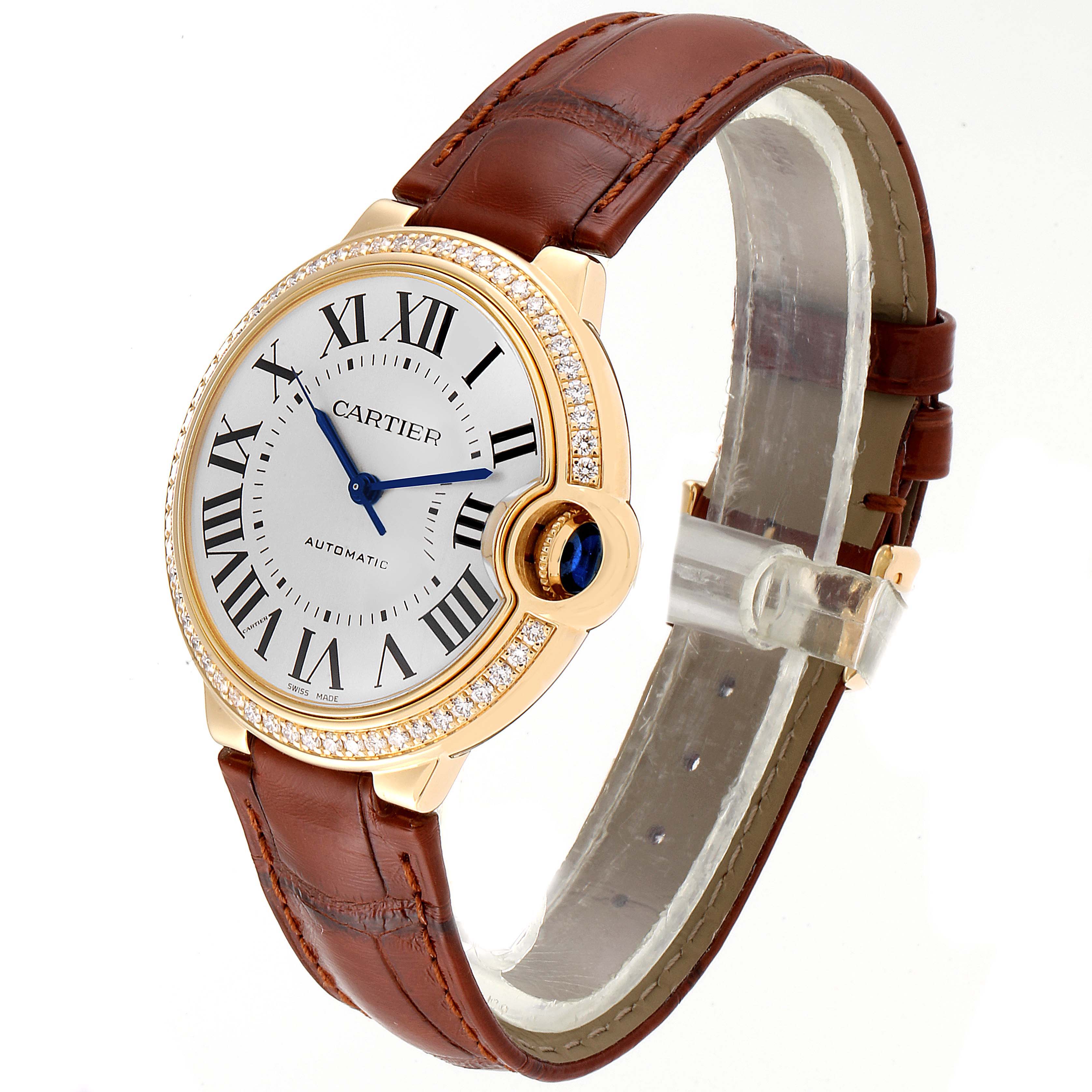The Cartier Ballon Bleu WE900451 Women
s Yellow Gold Silver Dial WE900451 Women
s Yellow Gold Silver Dial watch is shown at an angle displaying the face, Roman numerals, blue hands, diamond bezel, brown strap, and crown.