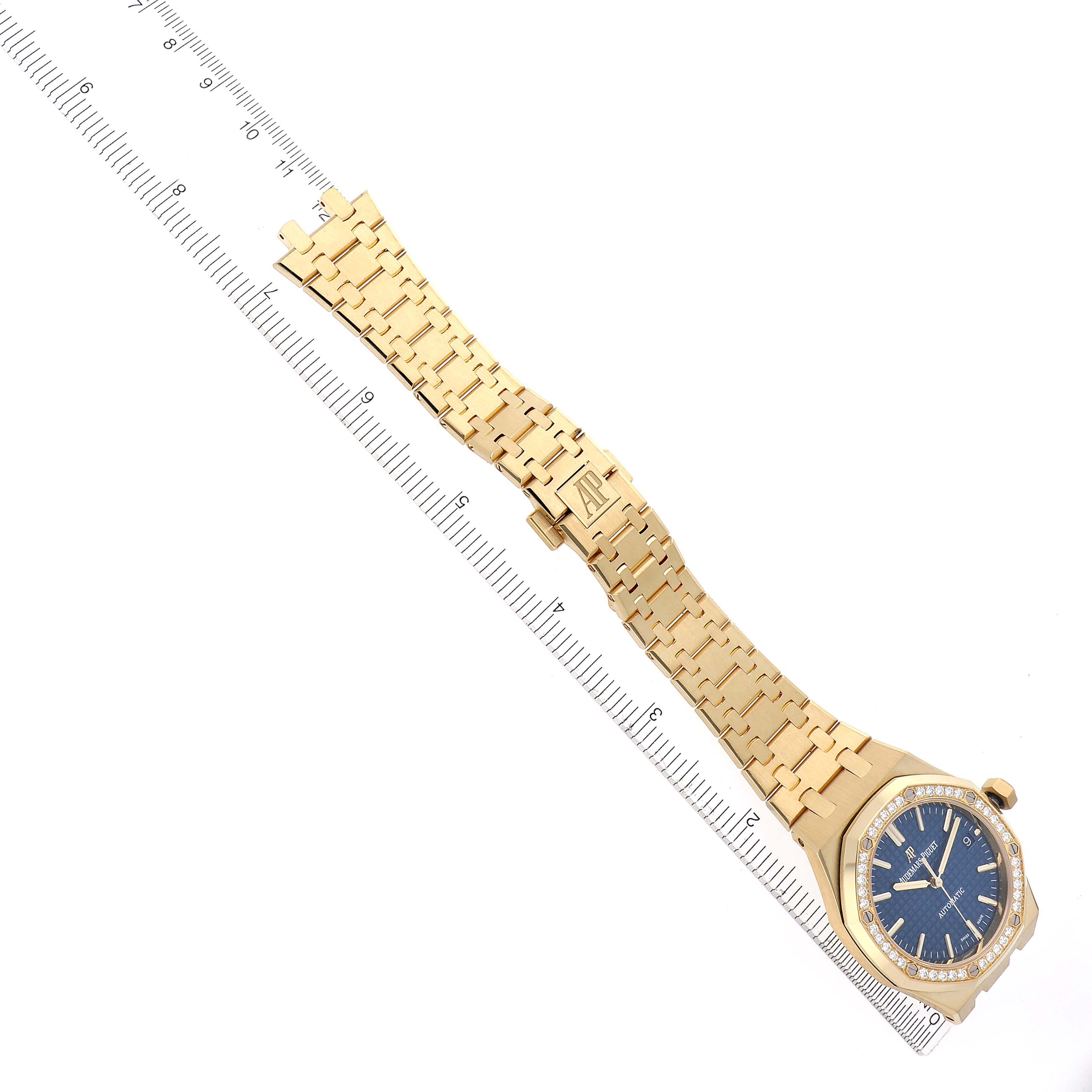 The image shows a top view of an Audemars Piguet Royal Oak 15451BA.ZZ.1256BA.01 Men's Yellow Gold Blue Dial 15451BA.ZZ.1256BA.01 Men's Yellow Gold Blue Dial watch, featuring its full bracelet, clasp, and face with a ruler for scale.