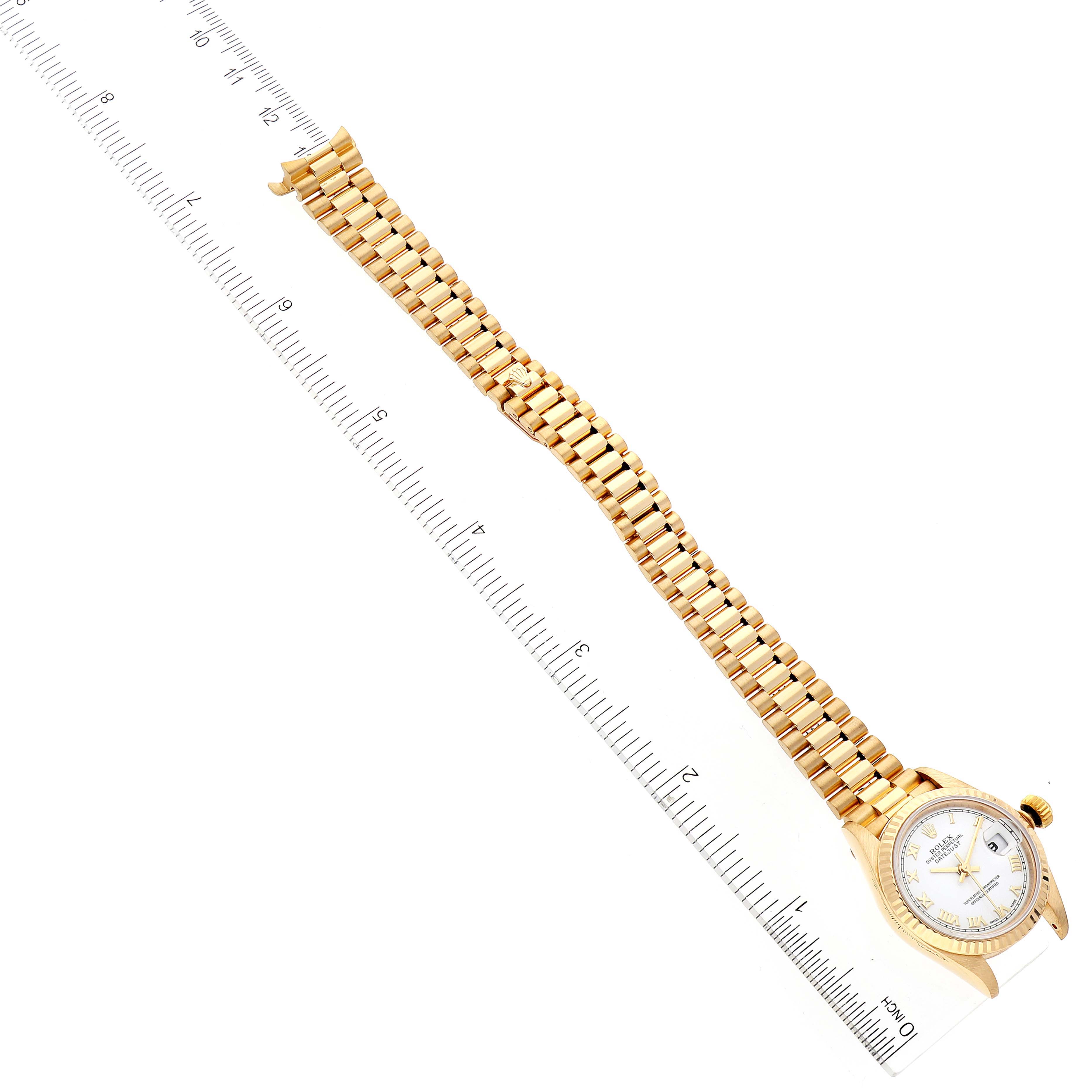The image shows a Rolex President 69178 Women
s Yellow Gold White Dial 69178 Women
s Yellow Gold White Dial watch from a top-down angle with its gold bracelet extended alongside a ruler.