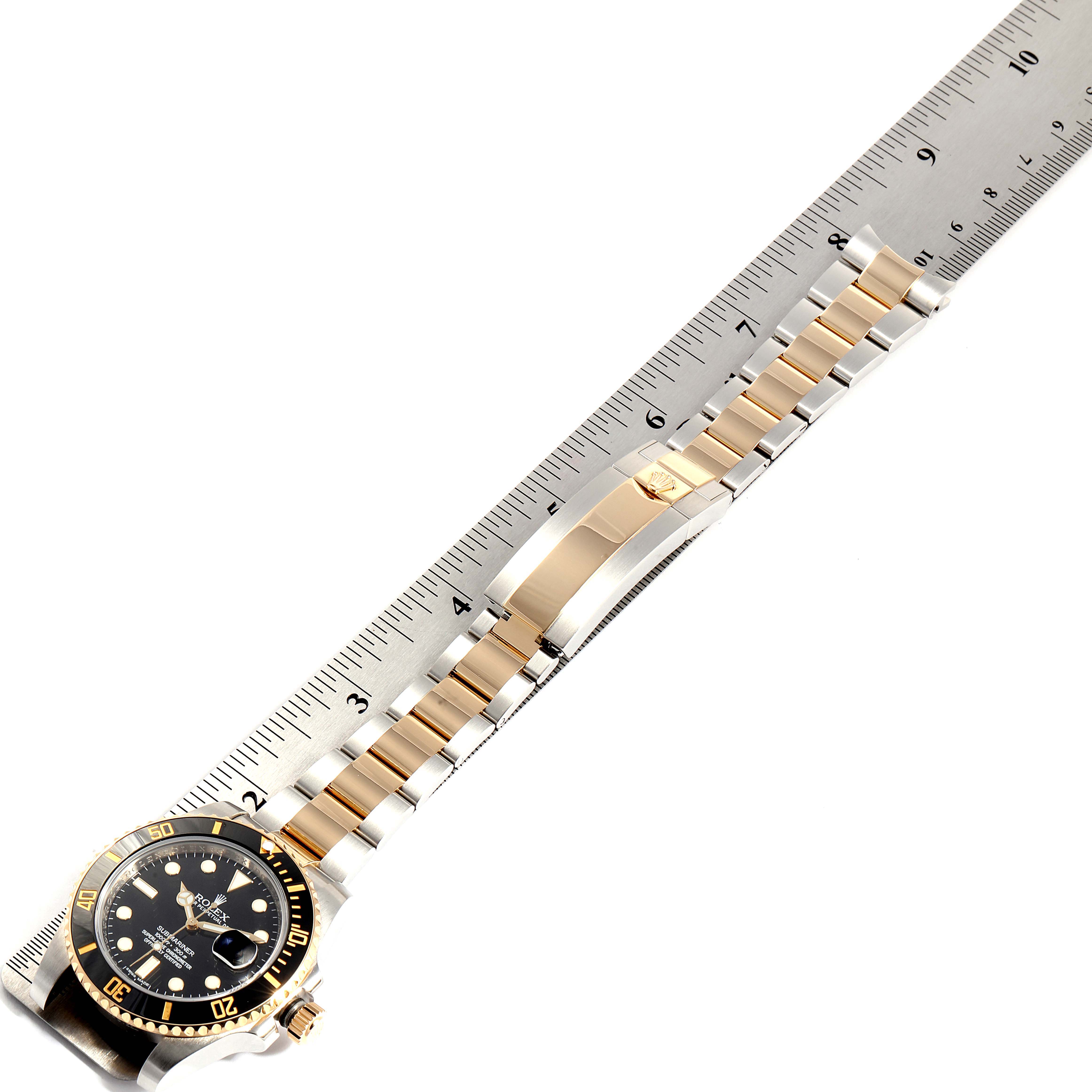 The image shows a Rolex Submariner 116613 Men
s Steel and Gold (two tone) Black Dial 116613 Men
s Steel and Gold (two tone) Black Dial watch laid flat with its gold and silver bracelet next to a ruler.