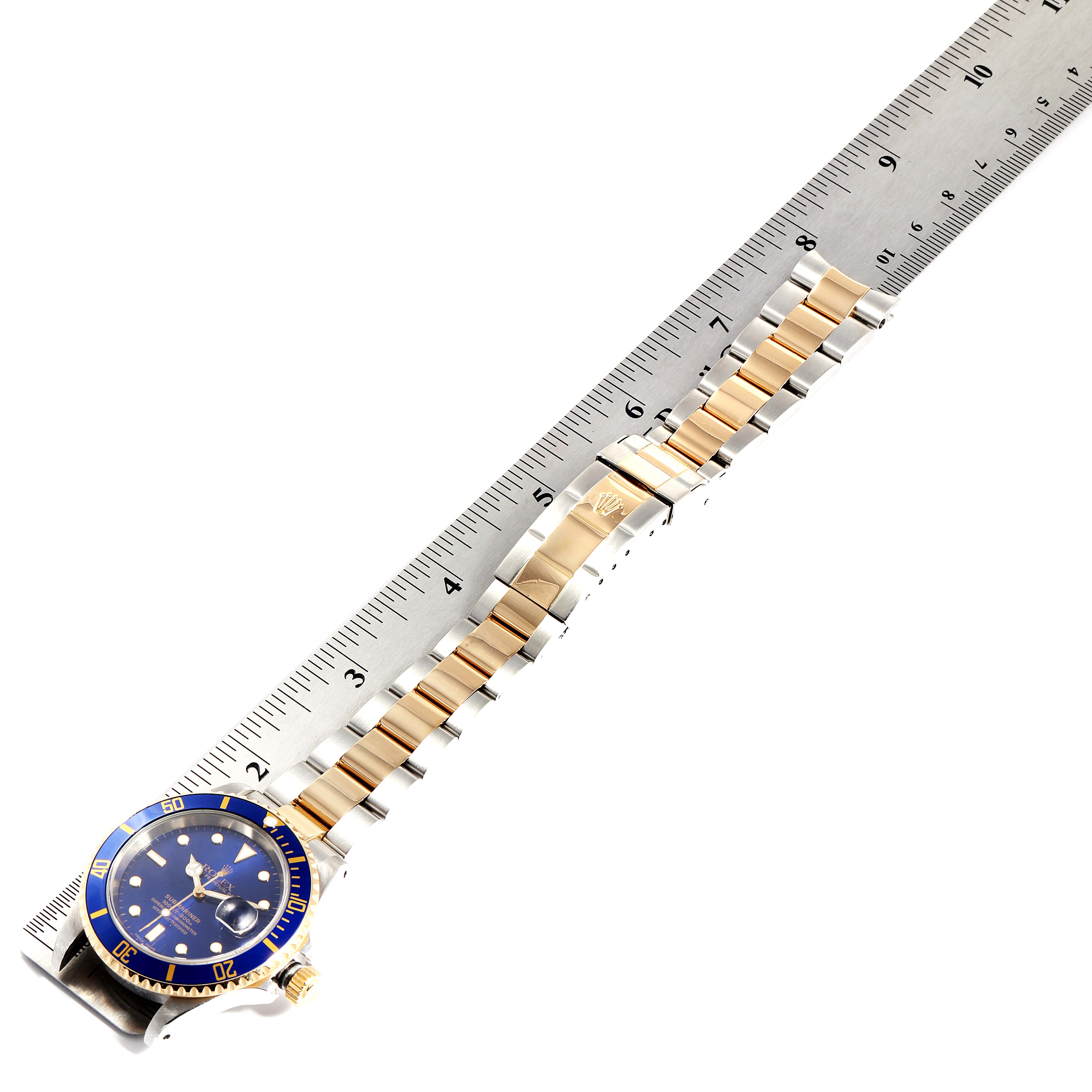 The image shows a Rolex Submariner 16613 Men
s Steel and Gold (two tone) Blue Dial 16613 Men
s Steel and Gold (two tone) Blue Dial watch placed alongside a ruler, highlighting its blue dial face, bezel, and two-tone bracelet.