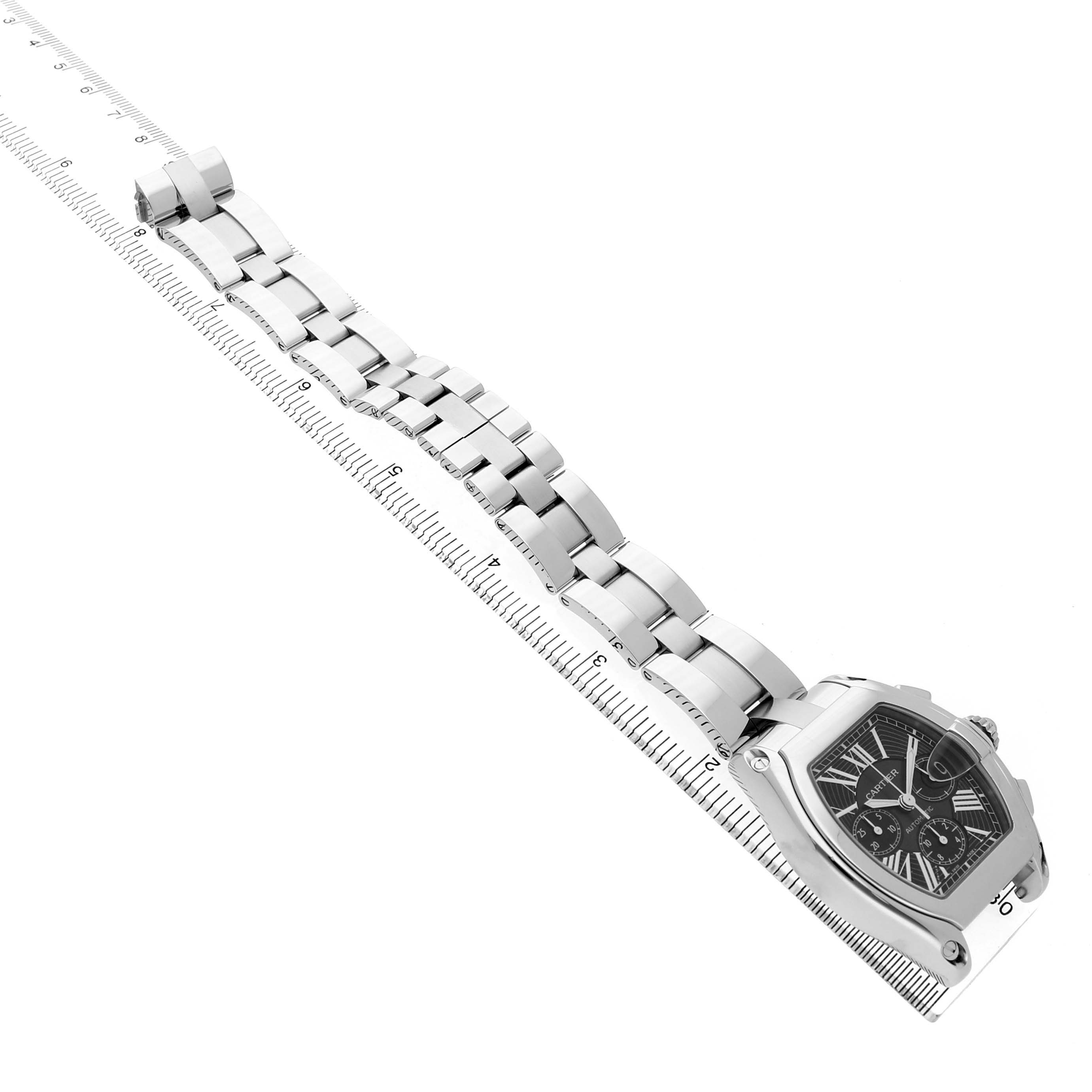The Cartier Roadster W62020X6 Men's Stainless Steel Black Dial watch is shown from a diagonal top-down angle, displaying its face and linked metal bracelet aligned with a ruler.