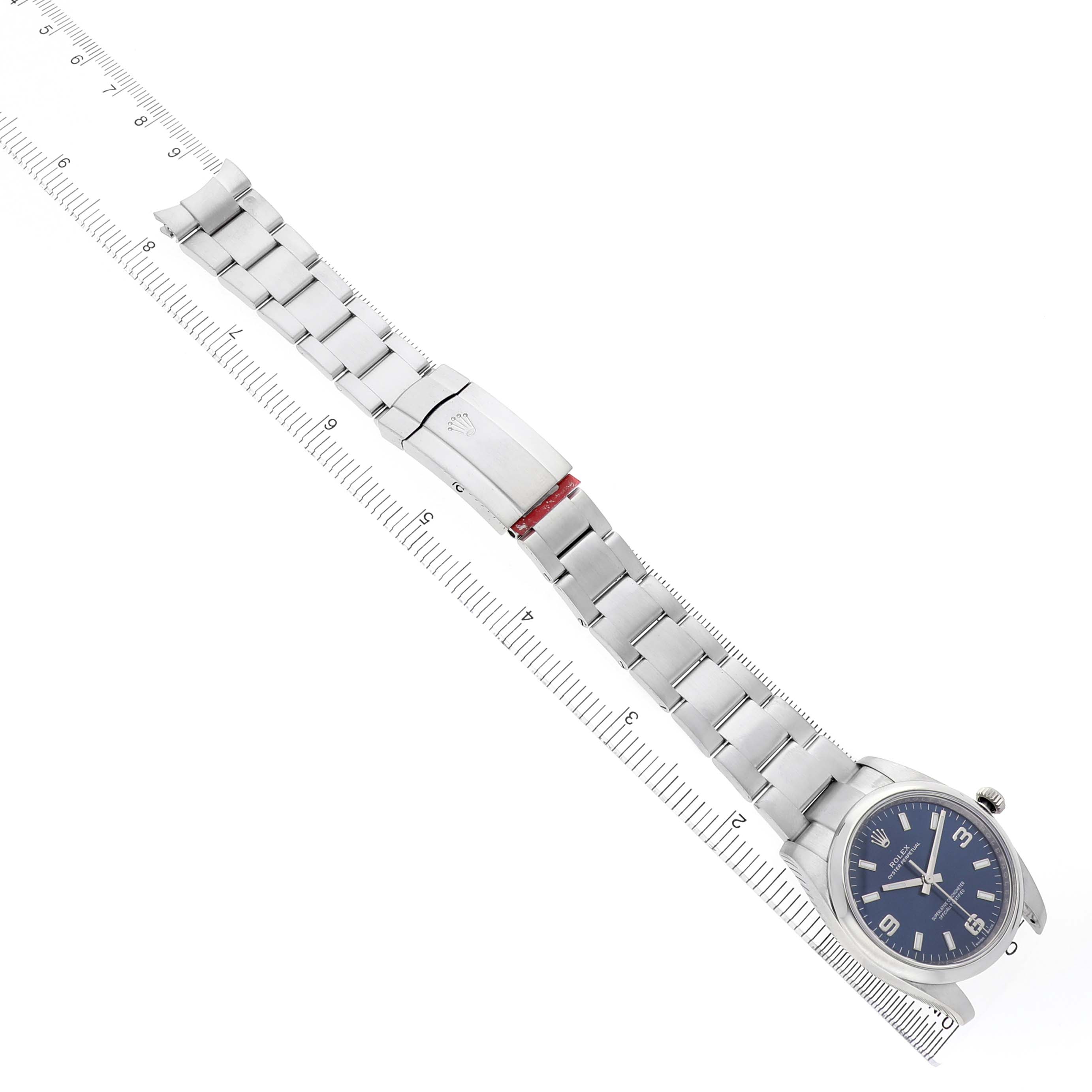 The Rolex Oyster Perpetual 114200 Men's Stainless Steel Blue Dial watch is shown from a top-down angle, displaying the bracelet, clasp, and face beside a ruler for scale.