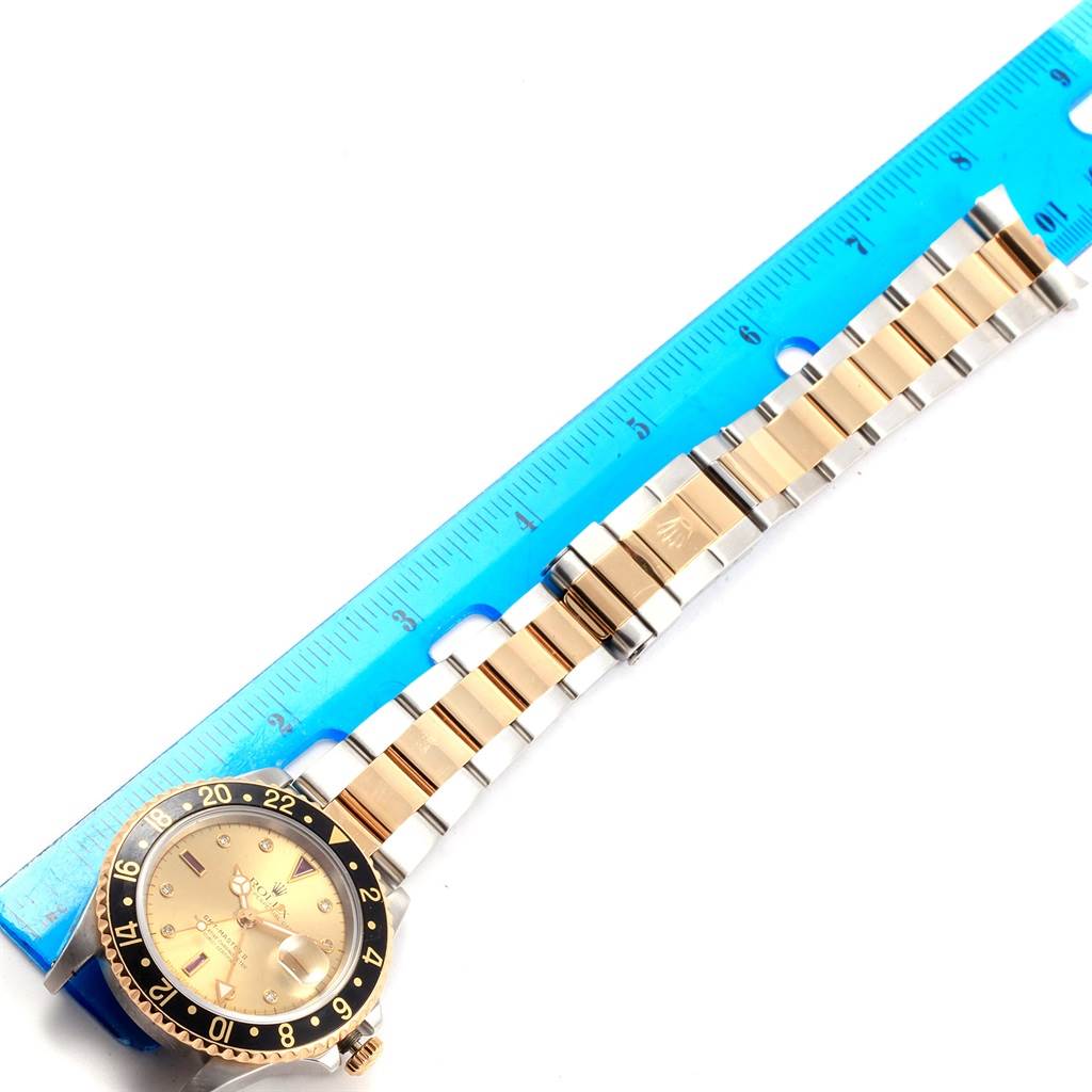 The image shows the front view of a Rolex 16713 Men
s Stainless Steel Champagne Dial 16713 Men
s Stainless Steel Champagne Dial GMT-Master II Yellow Gold Steel Diamond Ruby Serti Dial Watch 16713 with a blue ruler beside it.