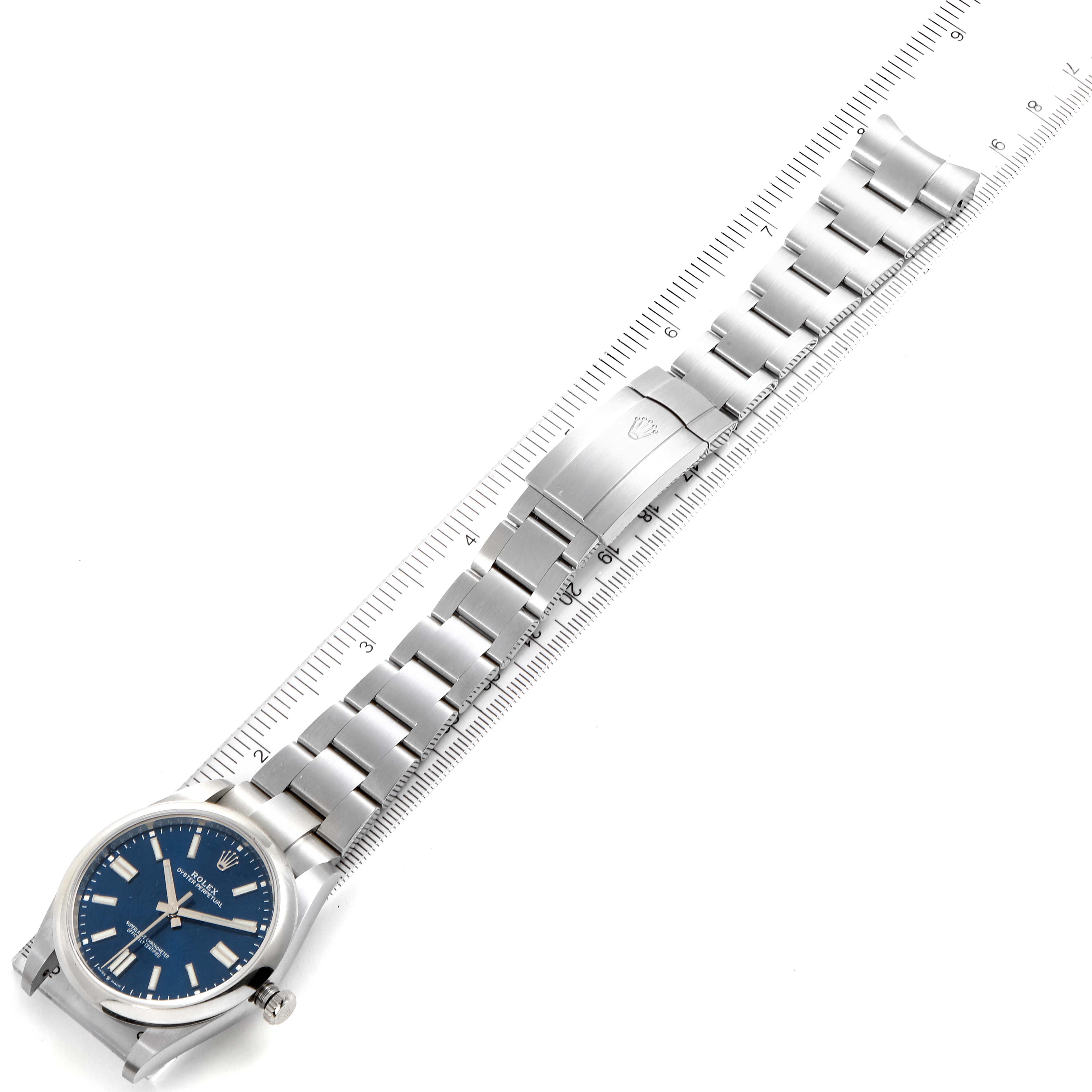 The image shows a complete overhead view of a Rolex Oyster Perpetual 114300 Men
s Stainless Steel Blue Dial 114300 Men
s Stainless Steel Blue Dial watch with a blue dial and stainless steel bracelet, next to a ruler.