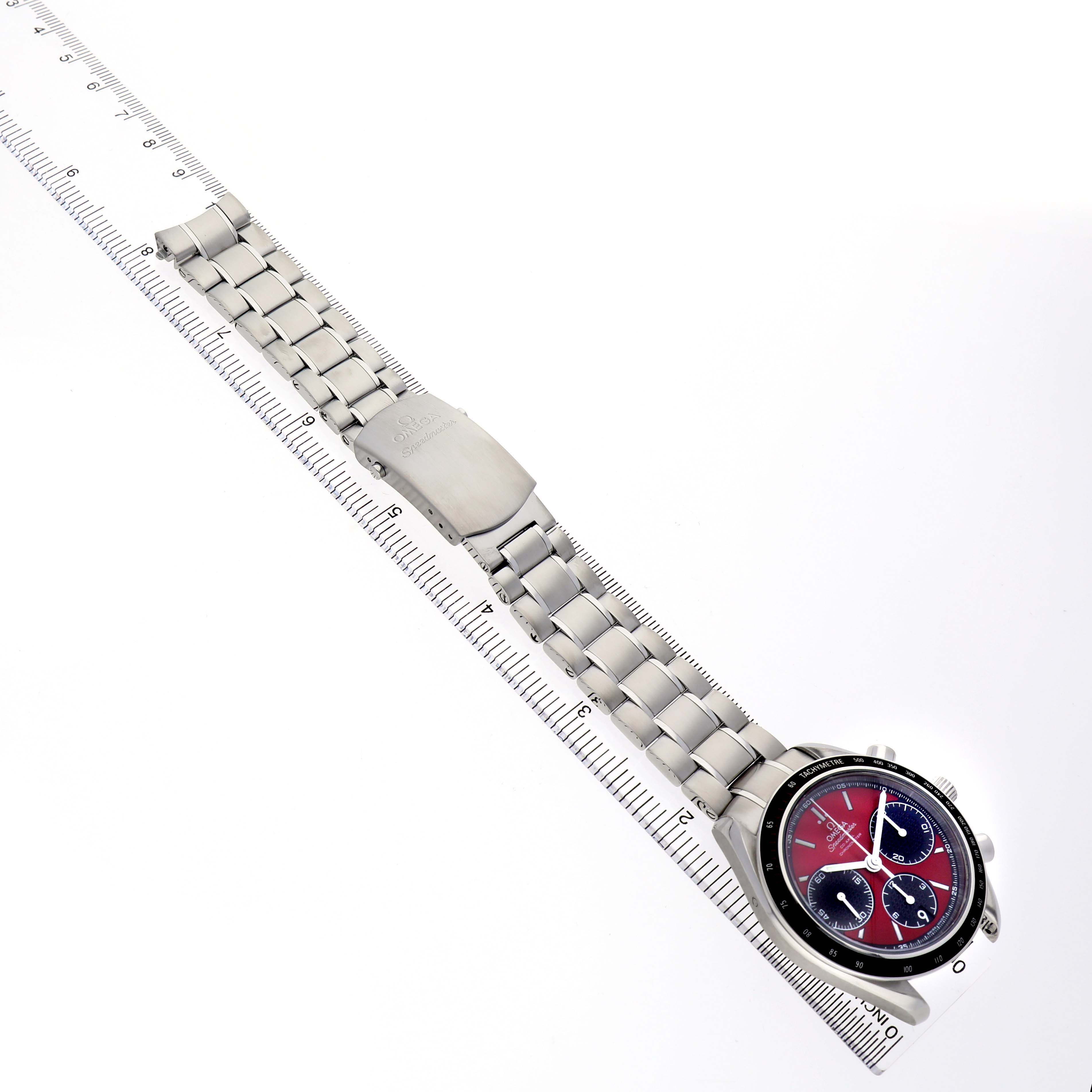 The Omega Speedmaster 326.30.40.50.11.001 Men's Stainless Steel Red Dial watch is shown from a top view, displaying its bracelet, clasp, and face alongside a ruler.