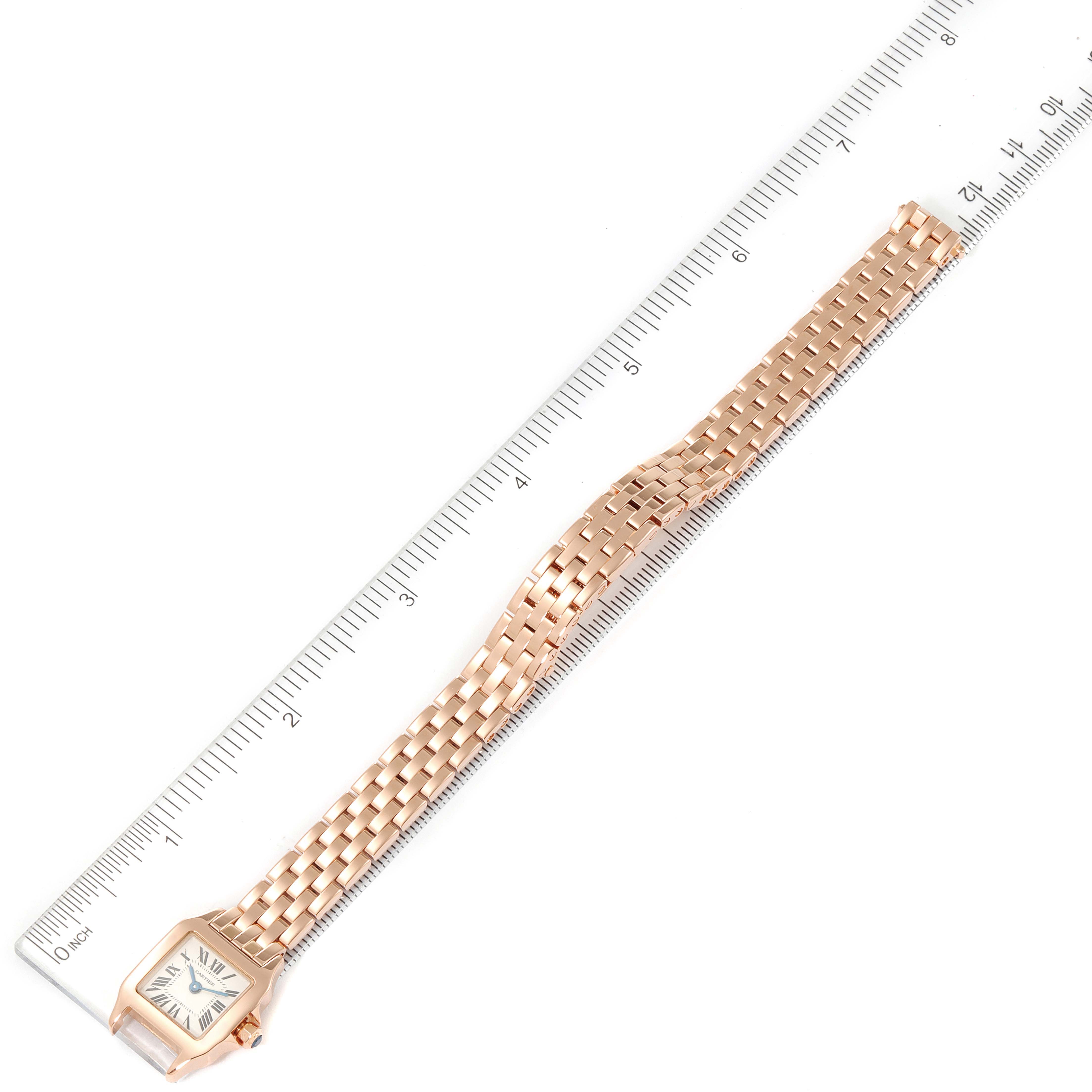 The image shows an overhead view of a Cartier Santos Demoiselle W25077X9 Women
s Rose Gold Silver Dial W25077X9 Women
s Rose Gold Silver Dial watch with its face and full gold bracelet next to a ruler for scale.
