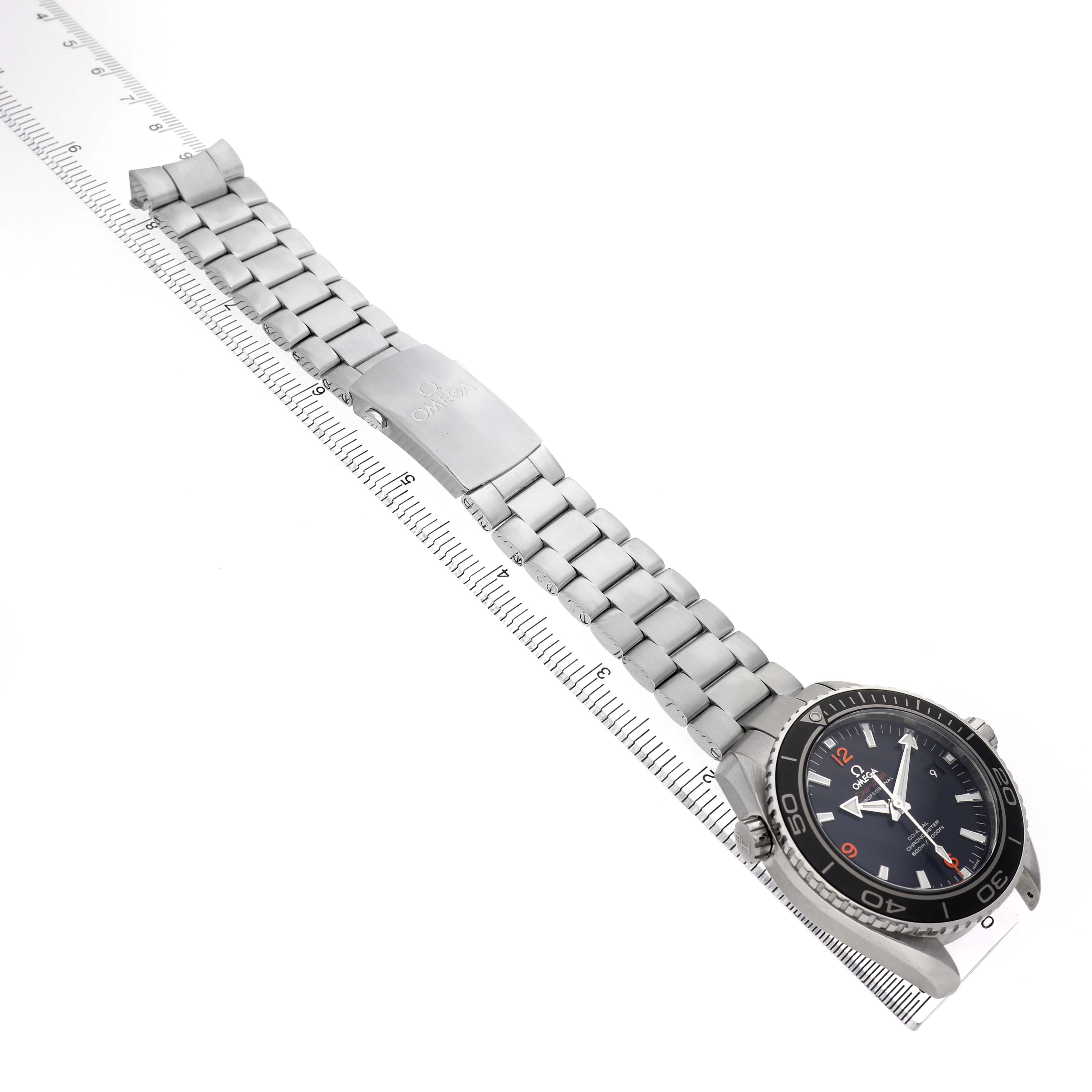 The Omega Planet Ocean 232.30.46.21.01.003 Men's Stainless Steel Black Dial watch is shown from a top-down angle, highlighting the face, bezel, and metal bracelet on a ruler.