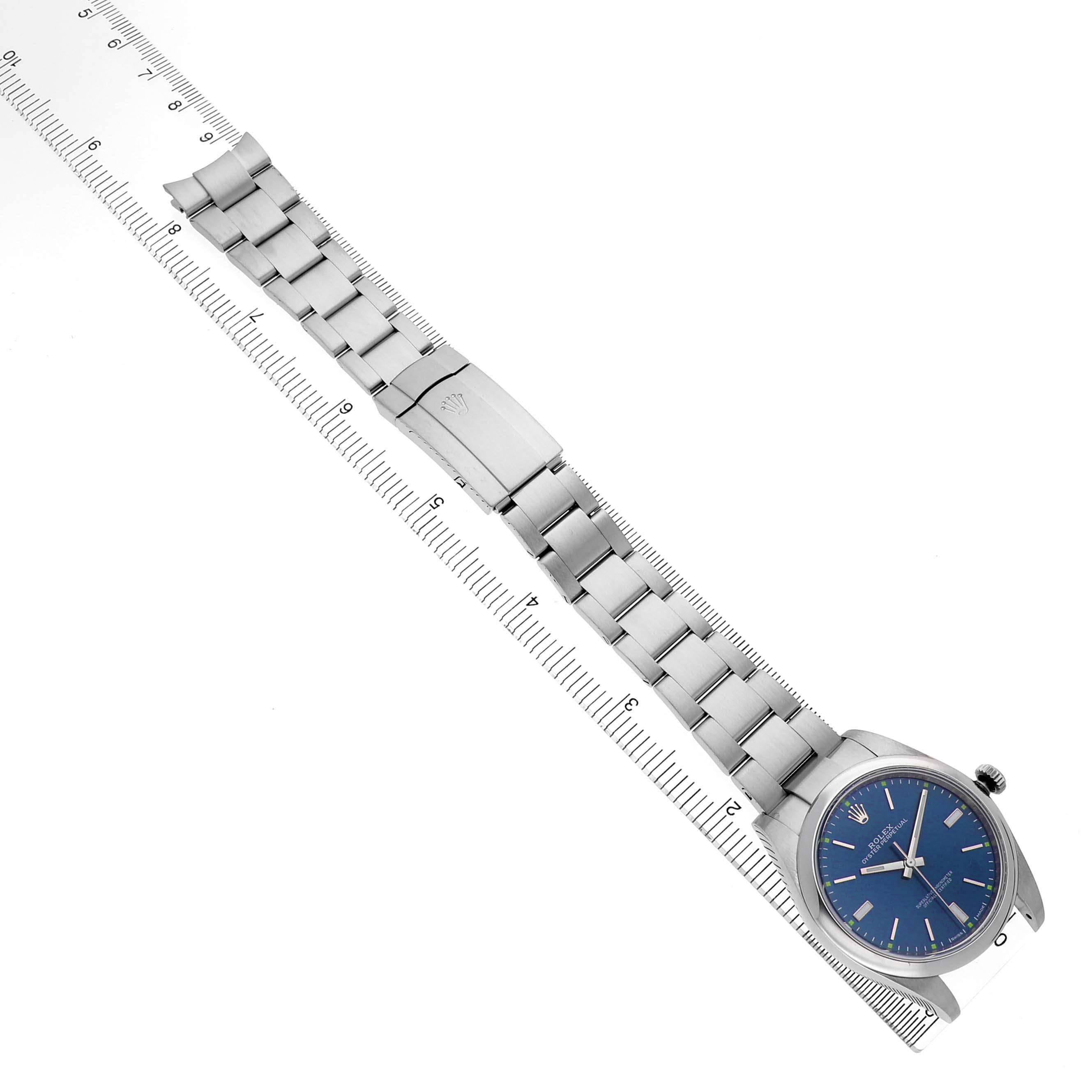 The Rolex Oyster Perpetual 114300 Men's Stainless Steel Blue Dial watch is shown from a top-down angle, displaying its blue dial, stainless steel bracelet, and clasp alongside a ruler.