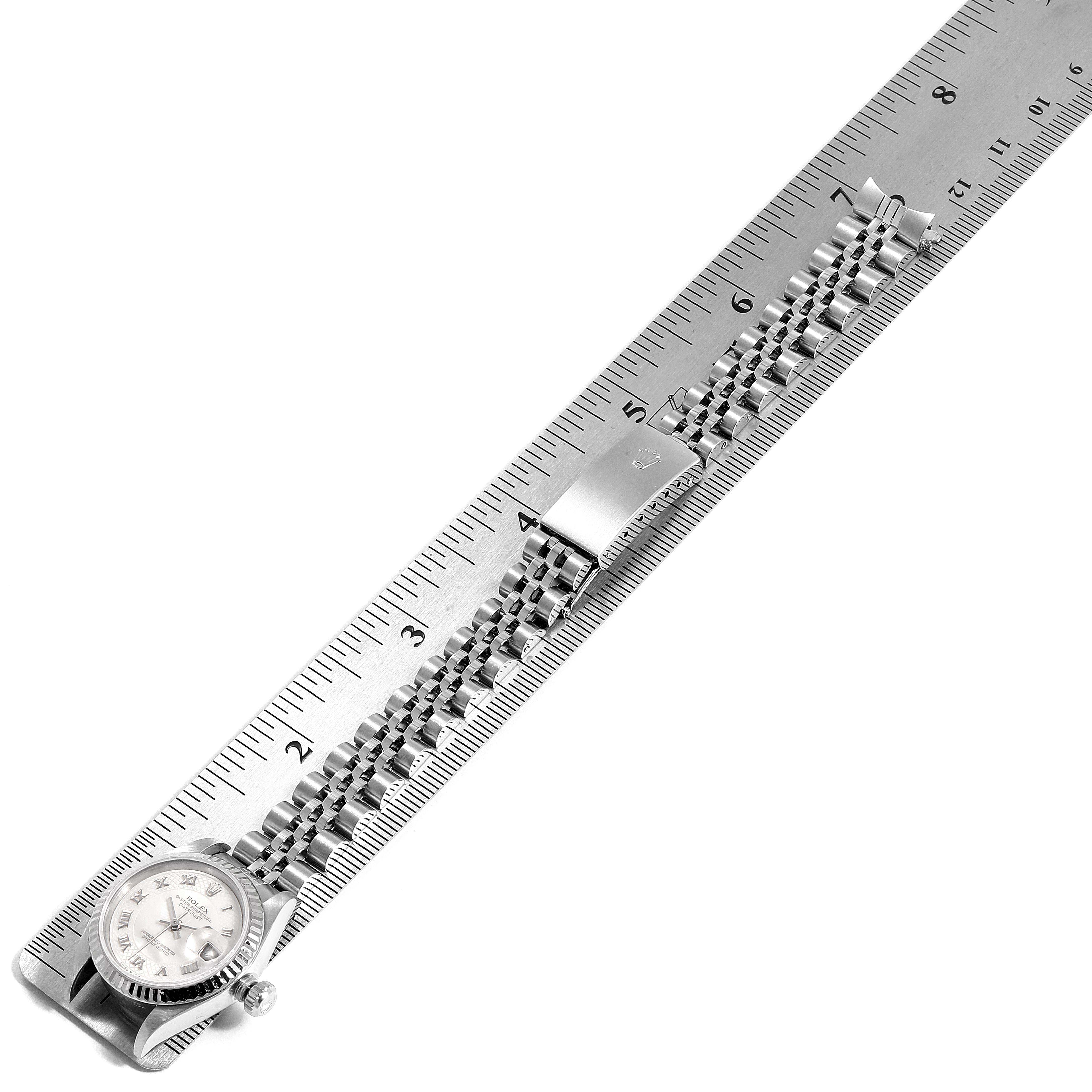 The image shows a Rolex Datejust 79174 Women
s Stainless Steel Mother of Pearl Dial 79174 Women
s Stainless Steel Mother of Pearl Dial watch laying flat with its band extended, alongside a ruler for size reference.