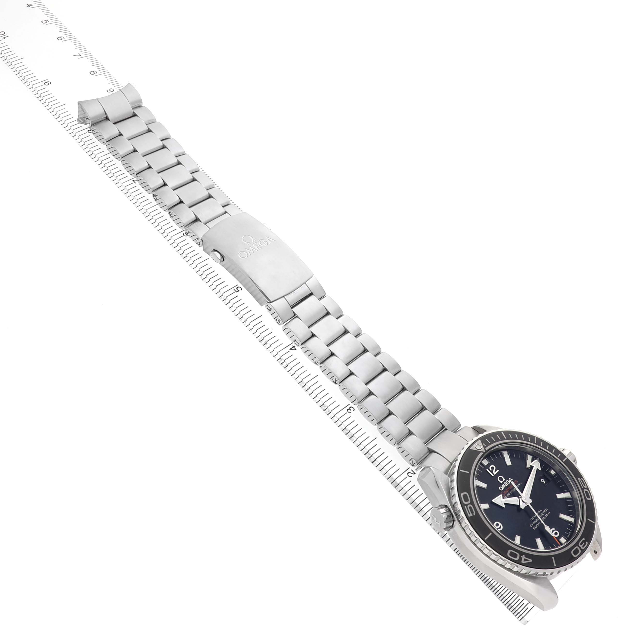 The Omega Planet Ocean 232.30.46.21.01.001 Men's Stainless Steel Black Dial watch is shown at a diagonal angle, displaying its face, bezel, and bracelet against a ruler backdrop.