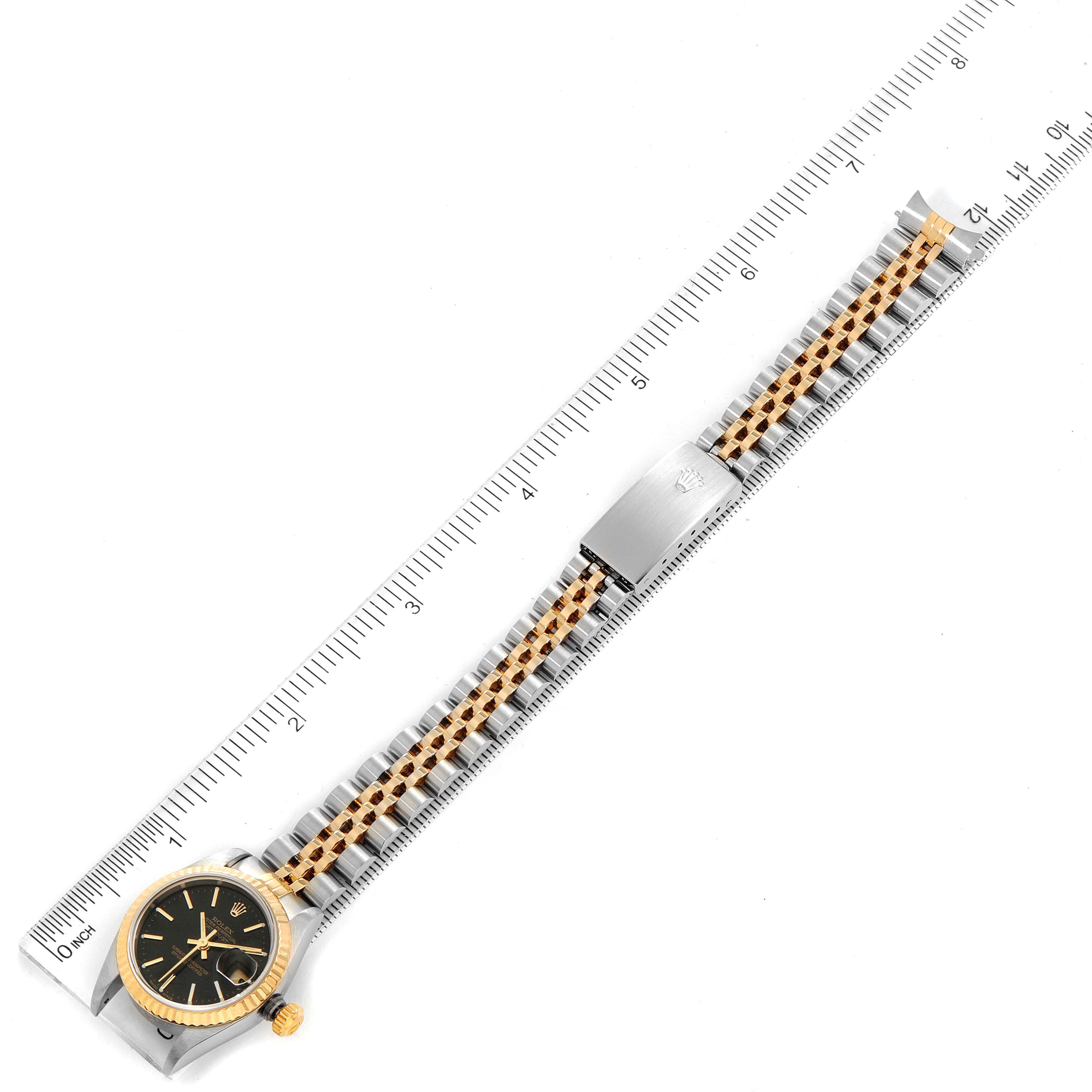 The image shows a top-down view of a Rolex Datejust 79173 Women
s Steel and Gold (two tone) Black Dial 79173 Women
s Steel and Gold (two tone) Black Dial watch, featuring the dial, case, and bracelet, placed against a ruler.