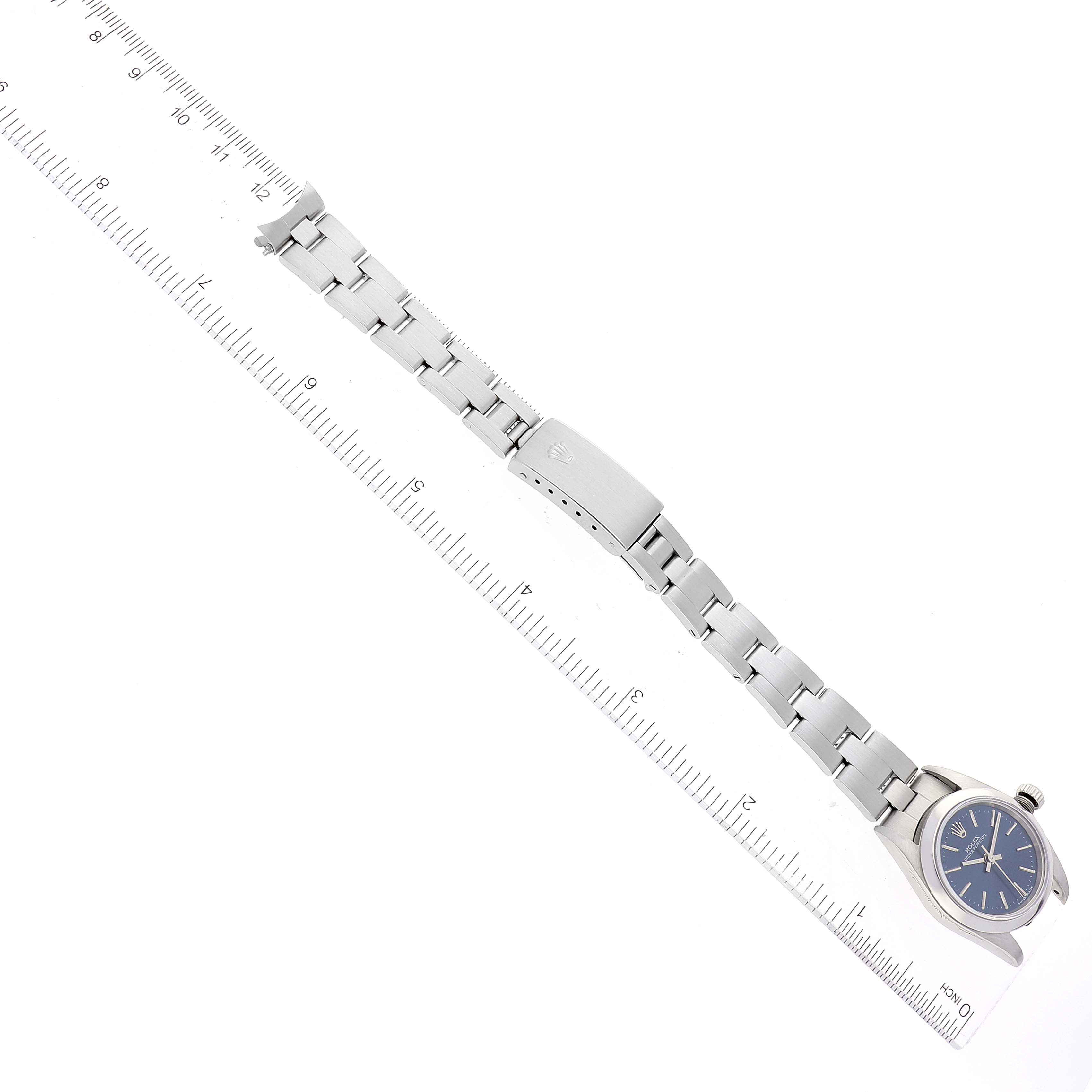 The image shows an overhead view of a Rolex Oyster Perpetual 76080 Women's Stainless Steel Blue Dial 76080 Women's Stainless Steel Blue Dial watch, including its face, bracelet, and clasp, alongside a ruler for scale.