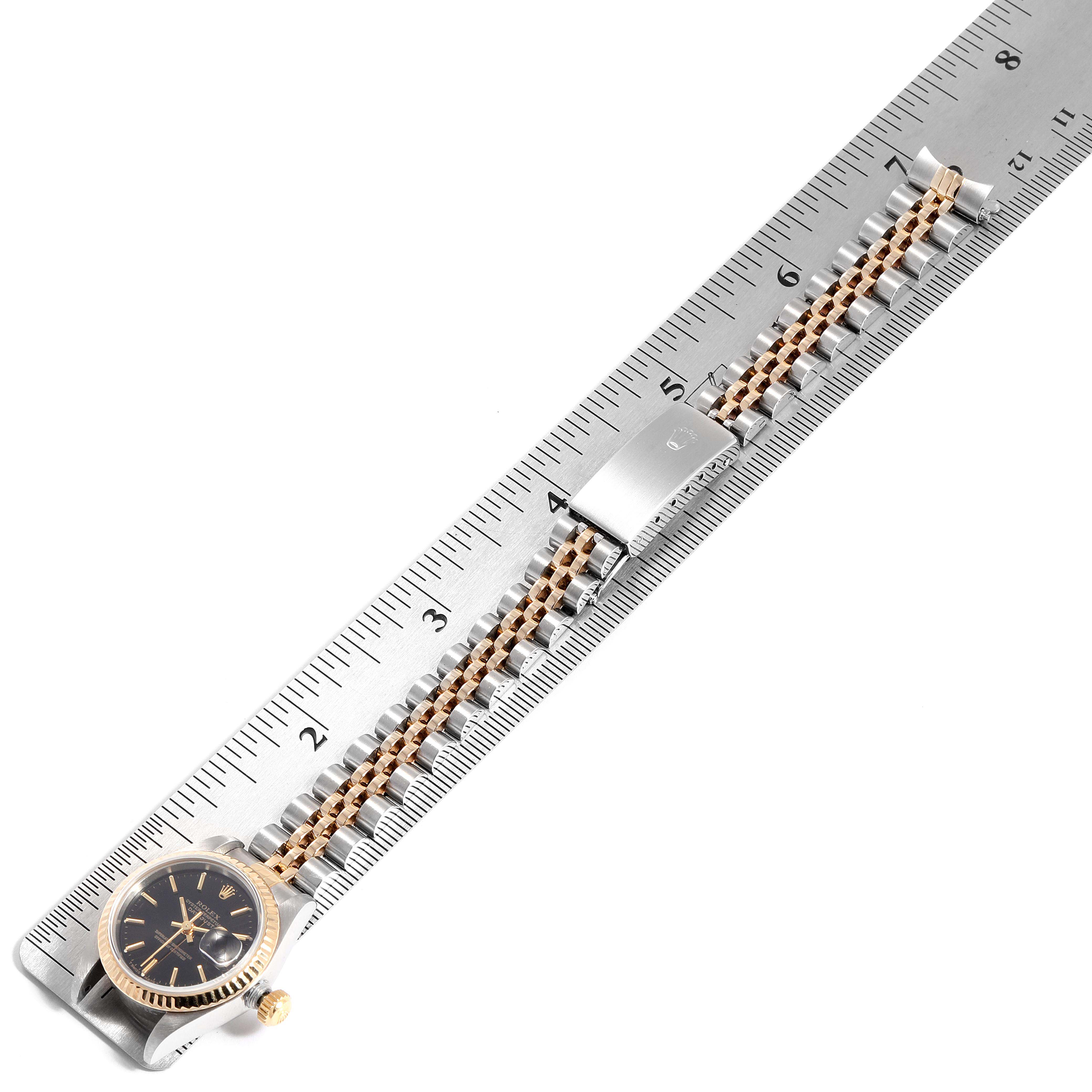 The picture shows a Rolex Datejust 69173 Women
s Steel and Gold (two tone) Black Dial 69173 Women
s Steel and Gold (two tone) Black Dial watch laid flat on a ruler, displaying its face, bracelet, and clasp.