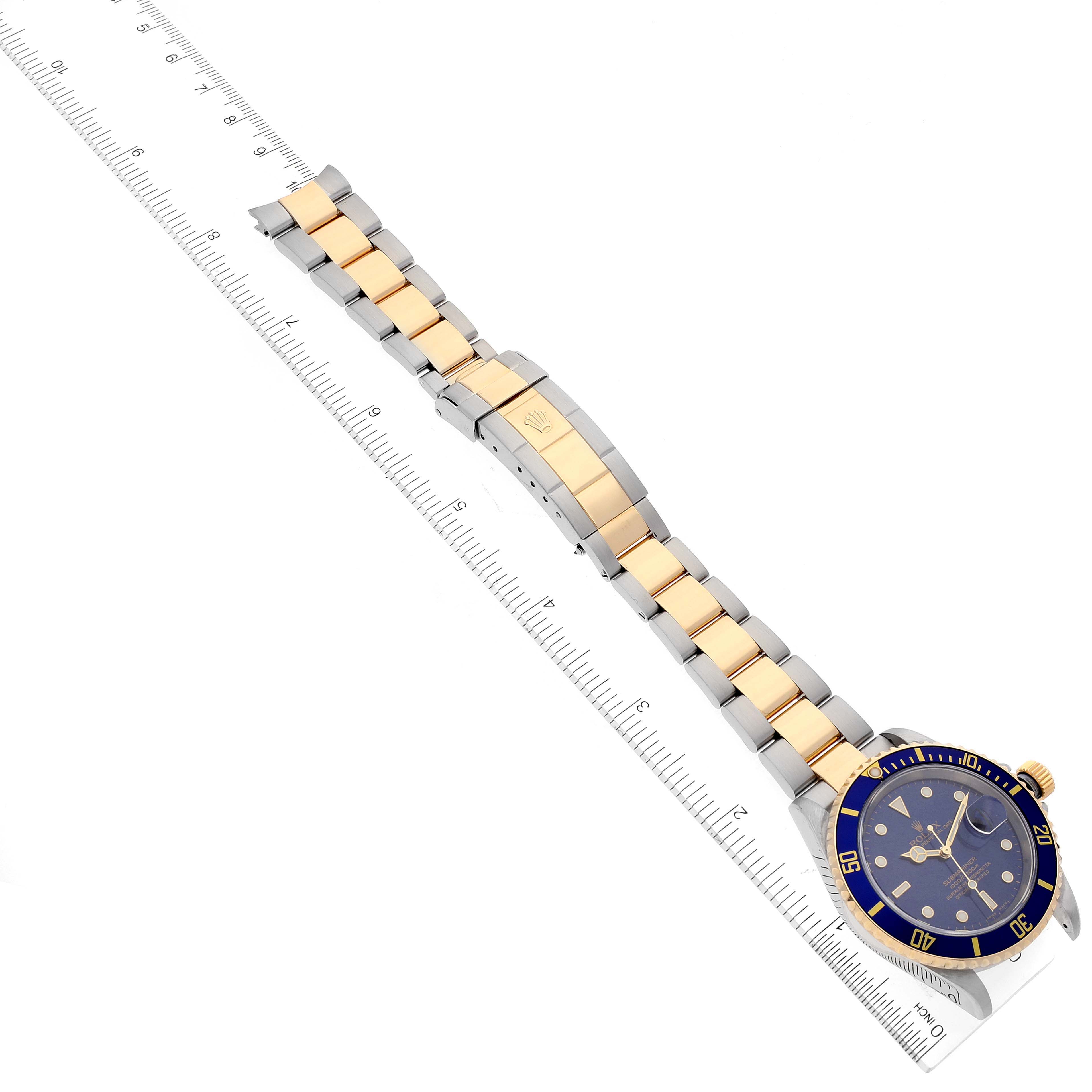 The image shows the top view of a Rolex Submariner 16613 Men
s Steel and Gold (two tone) Blue Dial 16613 Men
s Steel and Gold (two tone) Blue Dial watch with a two-tone bracelet placed on a ruler.