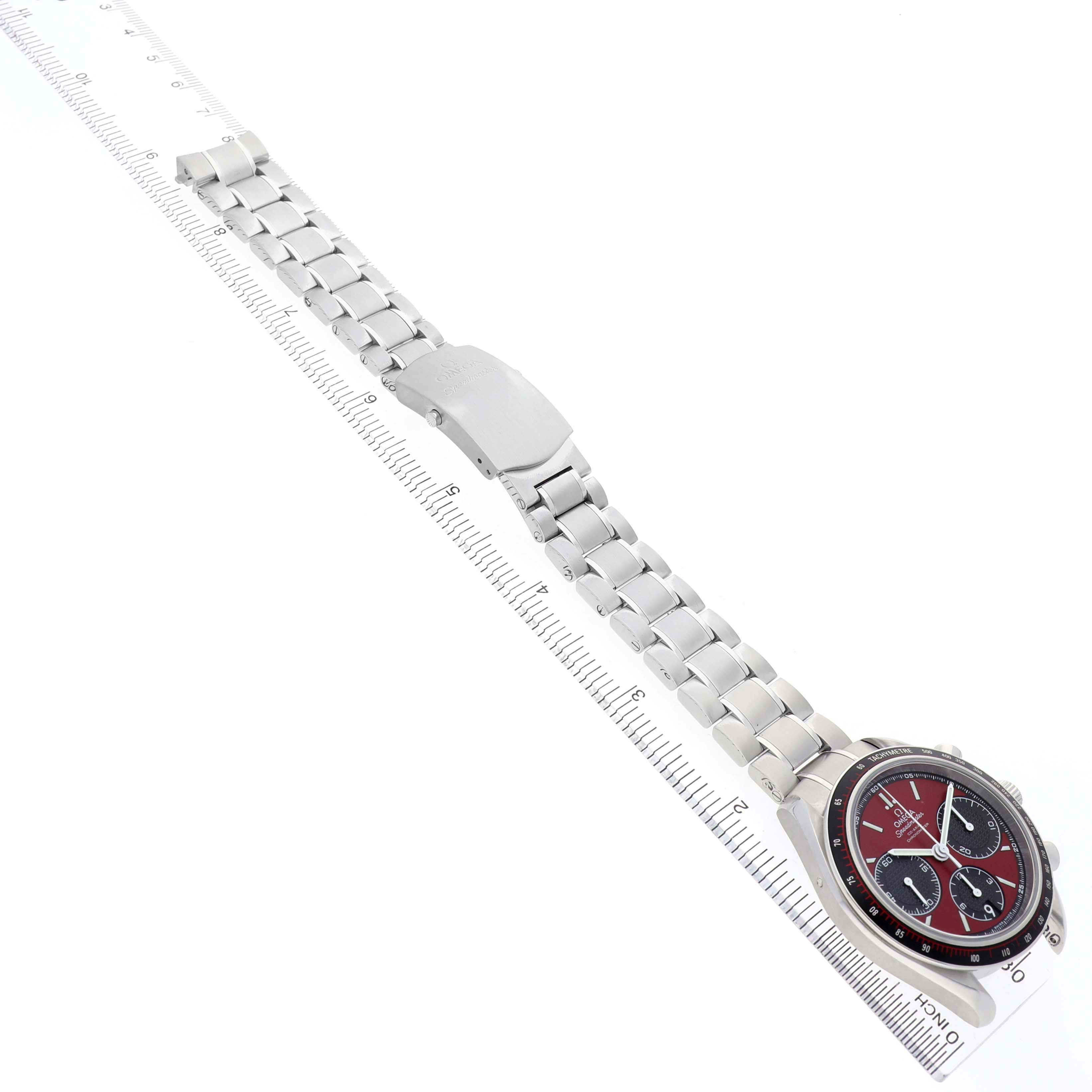 The Omega Speedmaster 326.30.40.50.11.001 Men's Stainless Steel Red Dial watch is shown from a top-down angle, highlighting the face, bracelet, and clasp on a ruler for scale.