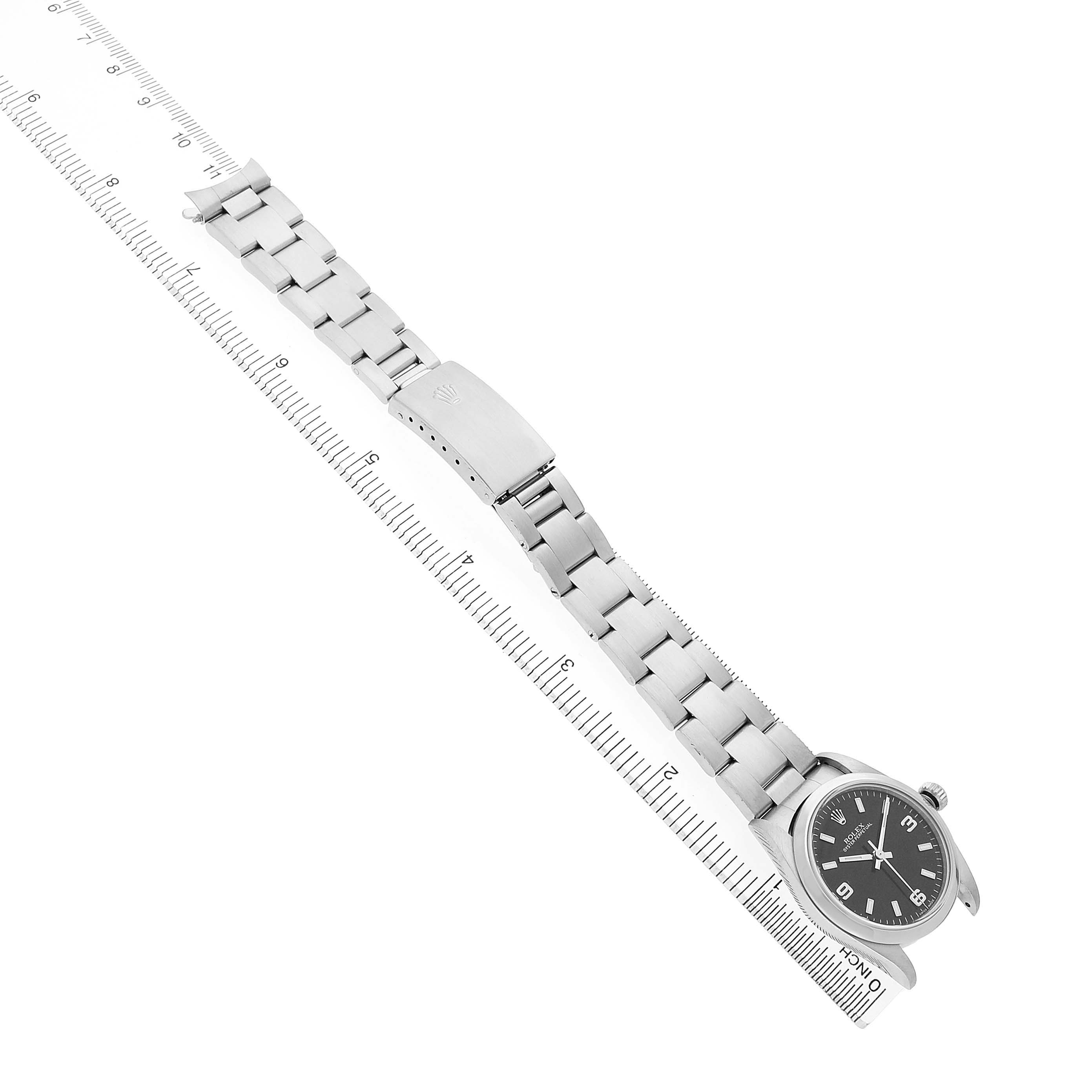 The Rolex Oyster Perpetual 77080 Women's Stainless Steel Black Dial watch is shown from an overhead angle, displaying the face, bracelet, and clasp alongside a ruler for scale.