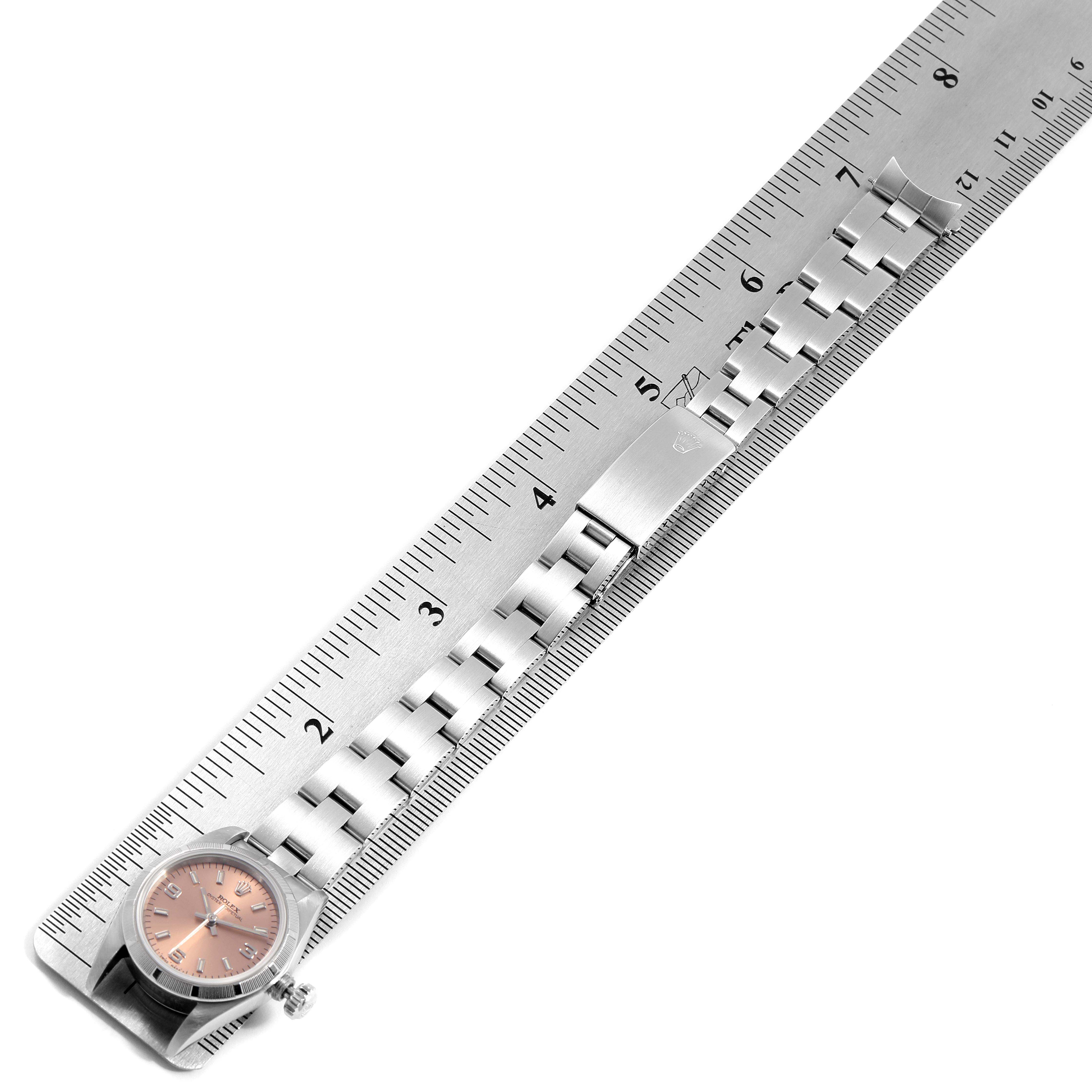 The image shows a side angle view of a Rolex Oyster Perpetual 67230 Women's Stainless Steel Pink Dial 67230 Women's Stainless Steel Pink Dial watch with its bracelet laid out alongside a ruler.