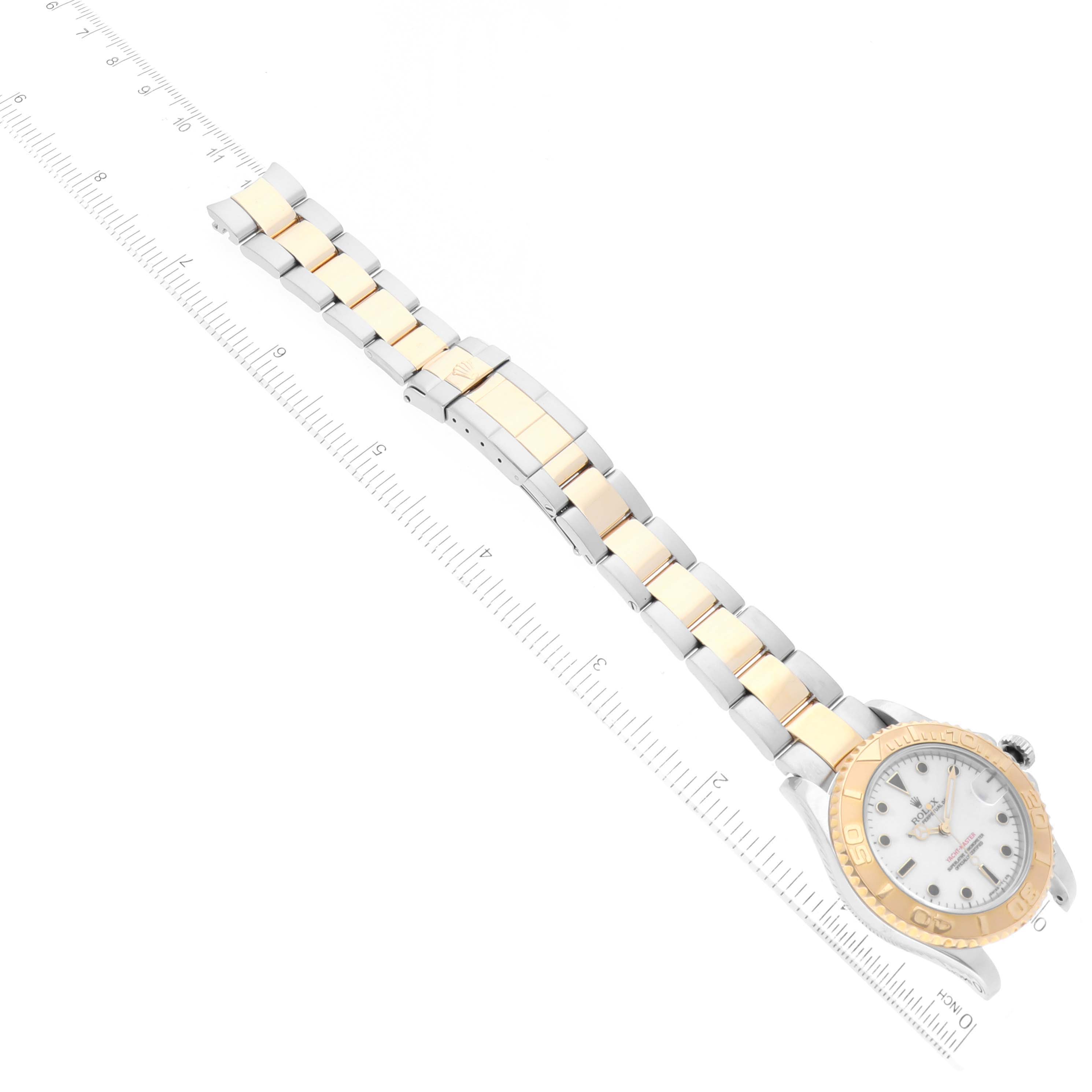 The image shows a Yacht-Master model Rolex watch laid flat with the face and gold and silver bracelet visible, alongside a ruler for scale.