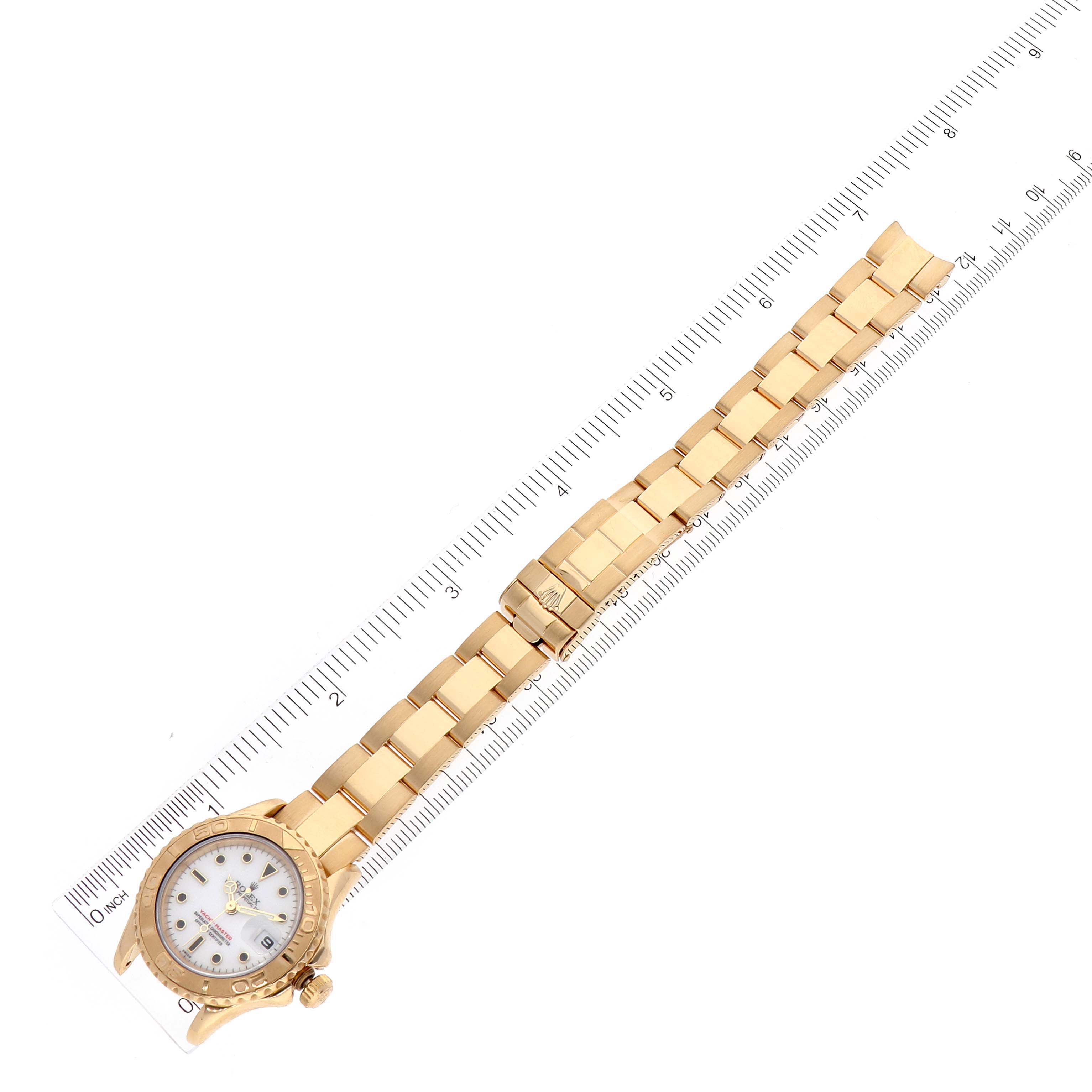 The Rolex Yacht-Master 69628 Women's Yellow Gold White Dial 69628 Women's Yellow Gold White Dial watch is positioned diagonally, showcasing its face and gold bracelet on a ruler for scale.