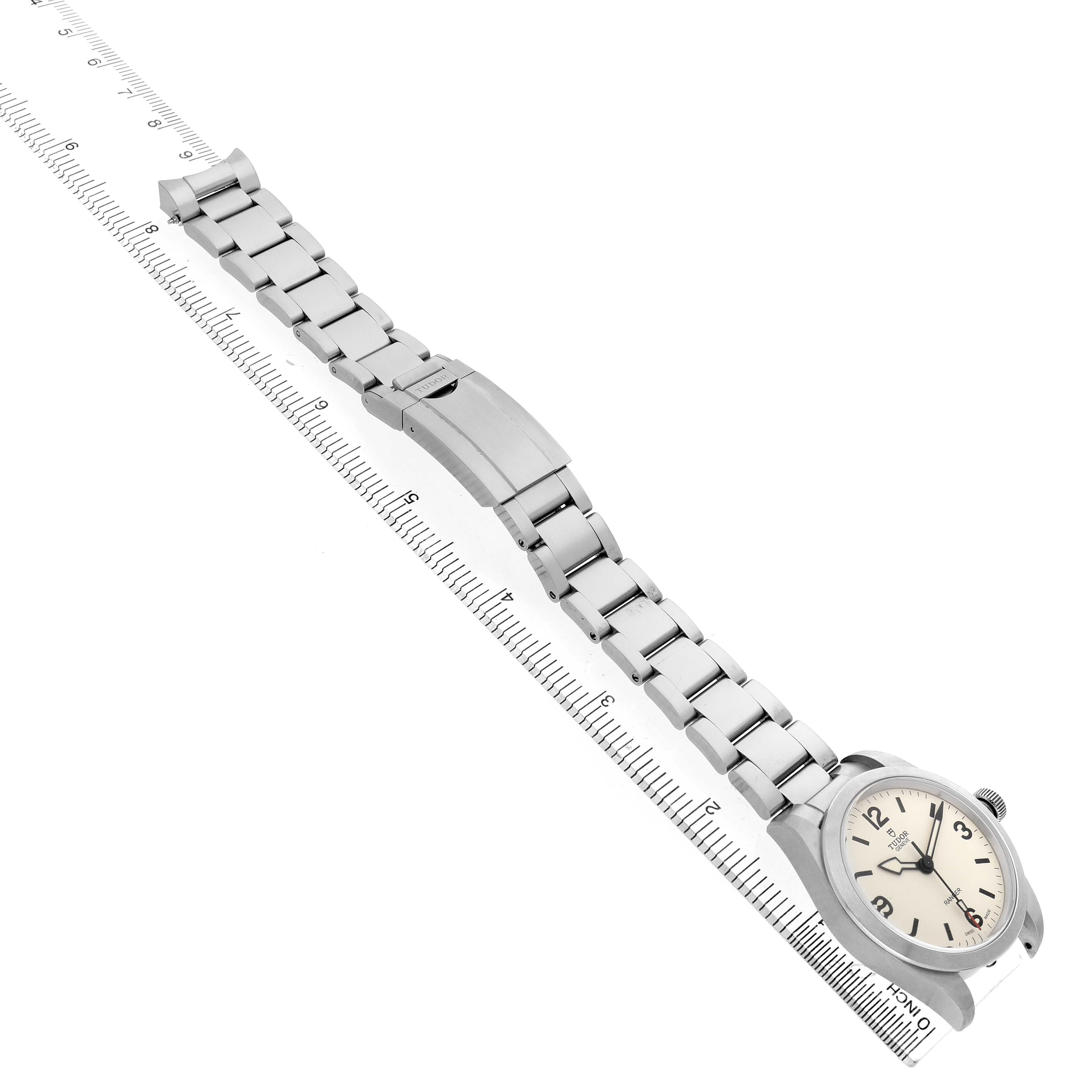 The Tudor Heritage Ranger 79930 Men's Stainless Steel Beige Dial watch is shown at an angle, highlighting the face, bracelet, and clasp against a ruler background.