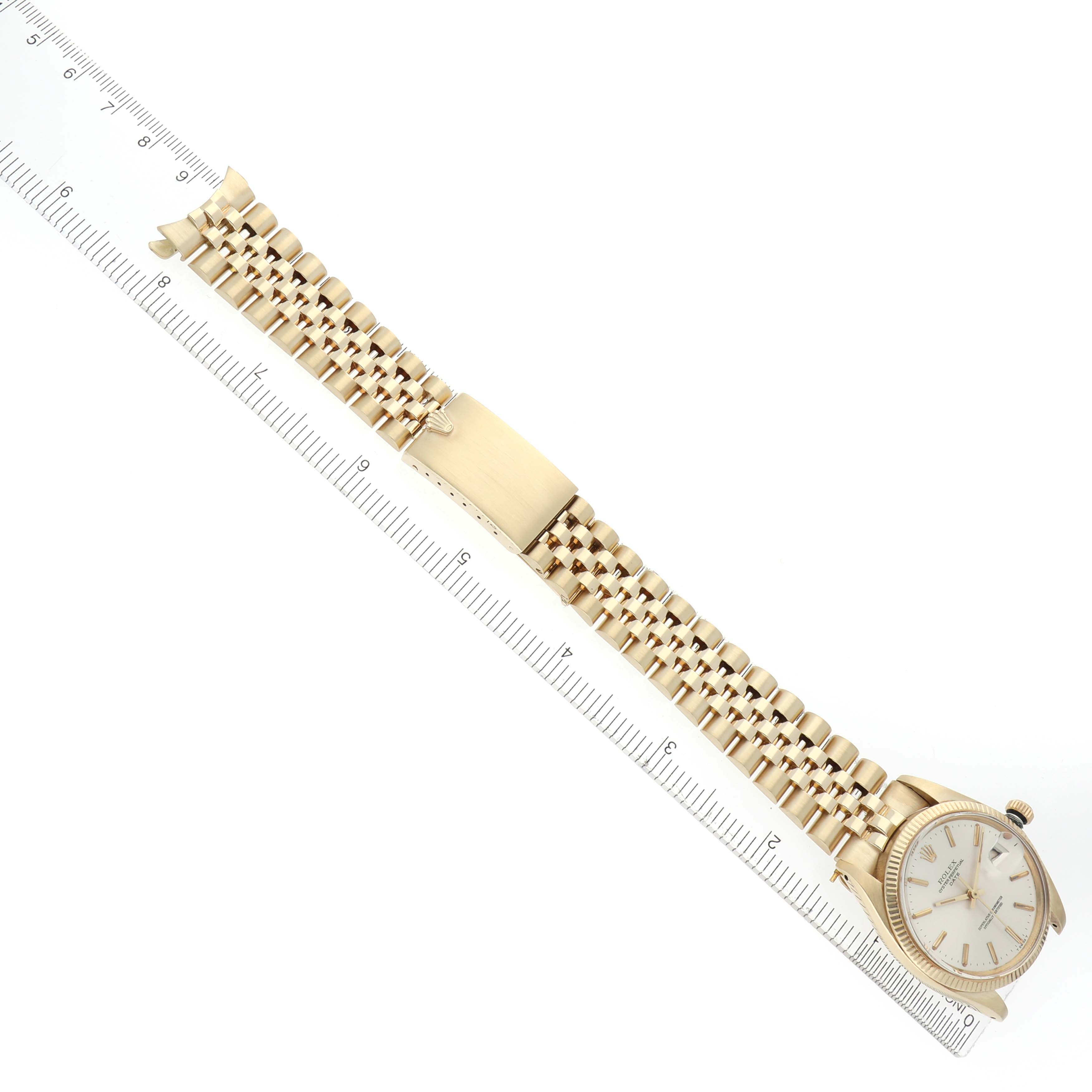 The Rolex Date watch is shown from a top view, displaying the dial, bracelet, and clasp alongside a measuring ruler.