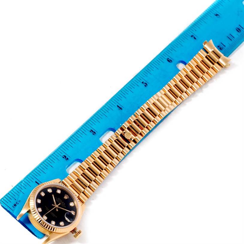The Rolex President 68278 Women's Yellow Gold Black Dial 68278 Women's Yellow Gold Black Dial watch is shown from the top angle with its full bracelet and face visible, next to a blue ruler.