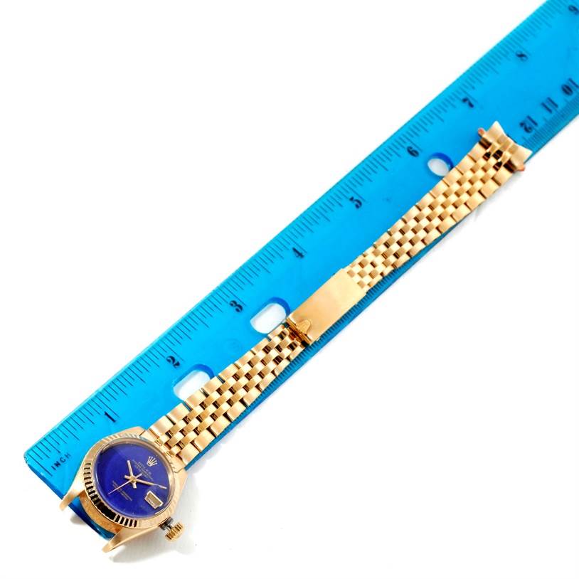 The Rolex President 6917 Women's Yellow Gold Blue Dial 6917 Women's Yellow Gold Blue Dial model watch is shown from a top-down angle, with its face and bracelet stretched out alongside a blue ruler.
