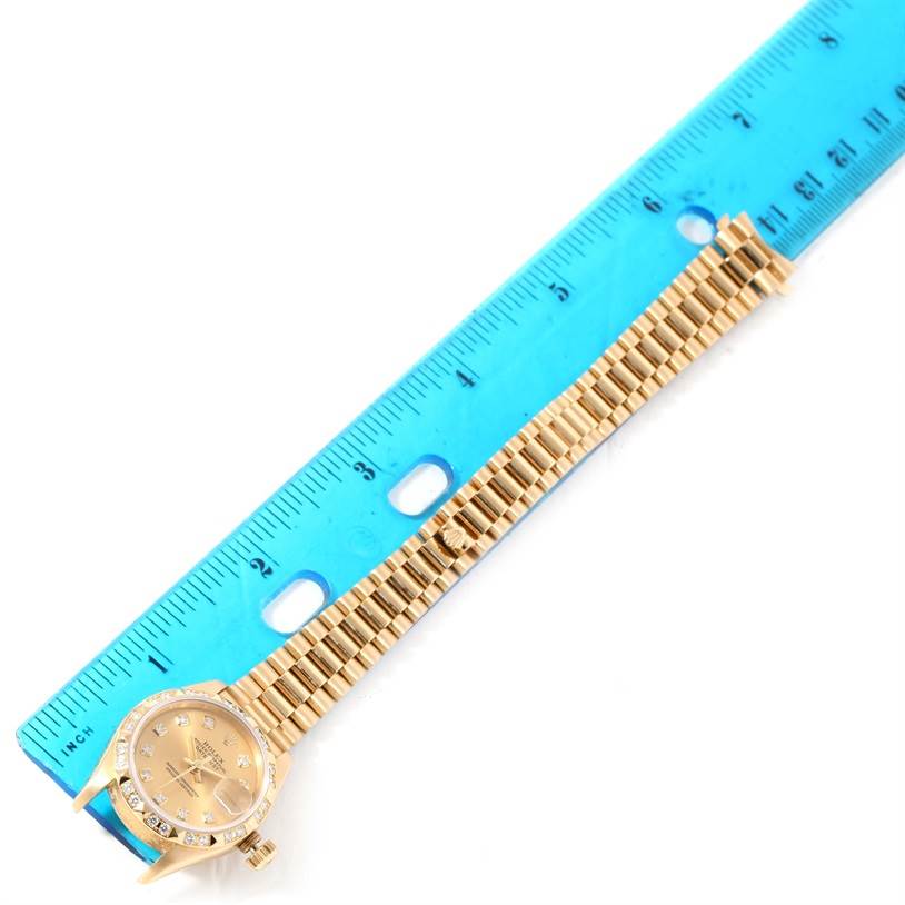The image shows a President model Rolex watch laid flat next to a blue ruler, displaying its face and bracelet.