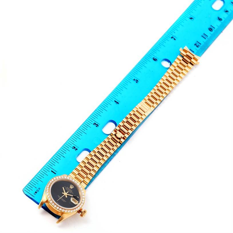 The image shows a Rolex President 69178 Women's Yellow Gold Black Dial 69178 Women's Yellow Gold Black Dial watch placed next to a blue ruler, highlighting the watch face, bezel, and bracelet.