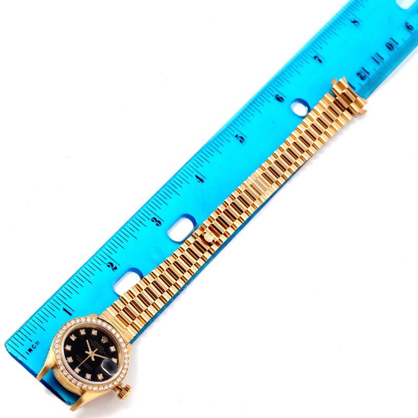 The Rolex President 69178 Women's Yellow Gold Black Dial 69178 Women's Yellow Gold Black Dial model watch is shown from a top-down angle, displaying the face, bezel, and yellow-gold bracelet next to a ruler.