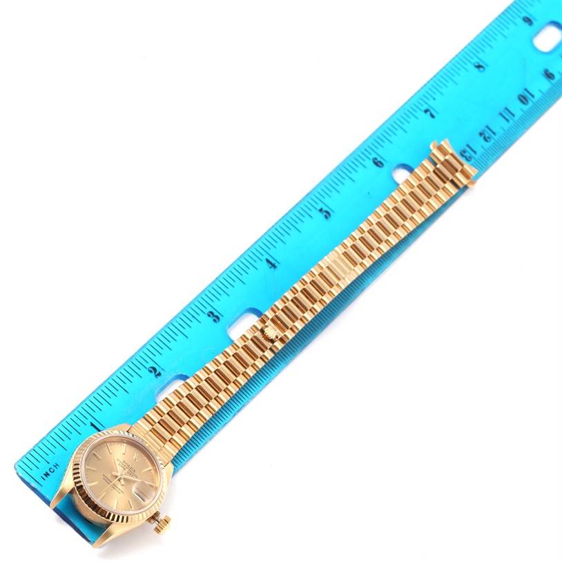 The image shows a Rolex President 69178 Women
s Yellow Gold Champagne Dial 69178 Women
s Yellow Gold Champagne Dial model watch with its bracelet aligned next to a blue ruler for measurement.