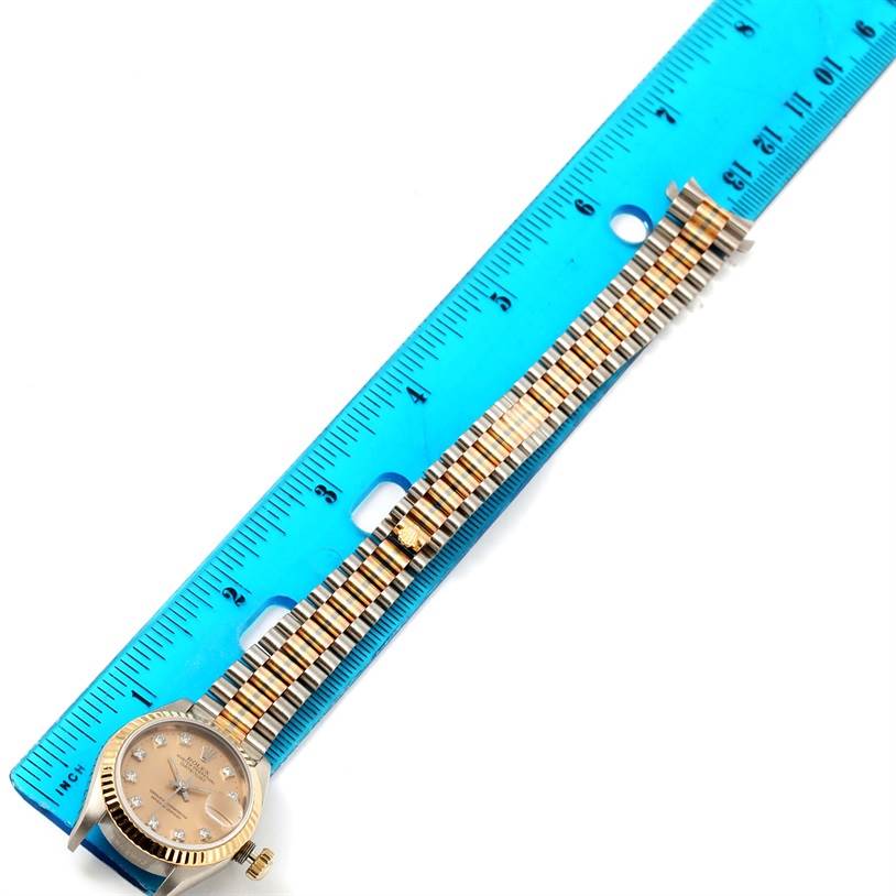 This image shows a Rolex President 69179 Women's White Gold Brown Dial 69179 Women's White Gold Brown Dial model watch with its band stretched out beside a blue ruler.