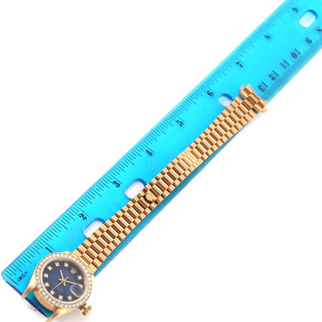 The image shows a Rolex President 69138 Women's Yellow Gold Blue Dial 69138 Women's Yellow Gold Blue Dial model watch with its gold bracelet fully extended next to a blue ruler.