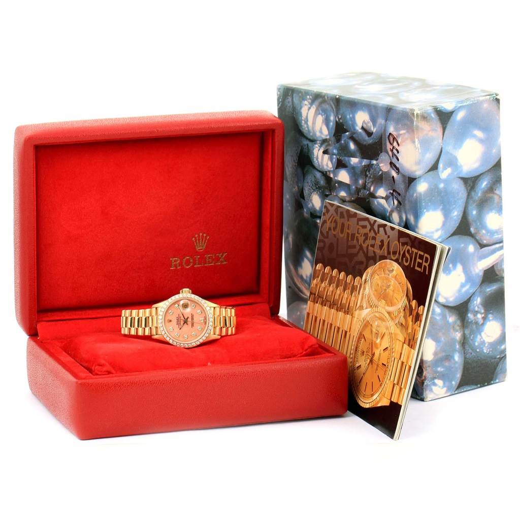 The Rolex President 69138 Women's Yellow Gold Pink Dial 69138 Women's Yellow Gold Pink Dial watch is displayed front-facing in a red box, with its manual and an outer packaging box to the side.