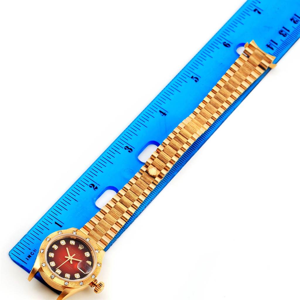 The image shows a Rolex President 79288 Women's Yellow Gold Red Dial 79288 Women's Yellow Gold Red Dial model watch, featuring its face and bracelet laid next to a blue ruler.