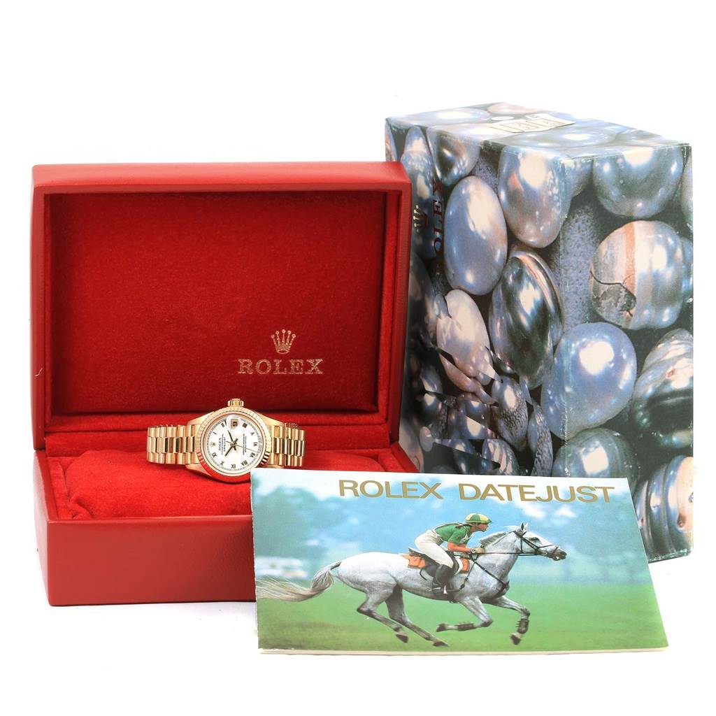 A Rolex President 79258 Women's Yellow Gold White Dial 79258 Women's Yellow Gold White Dial model watch is displayed in a red box, alongside its outer box and a Rolex Datejust booklet.