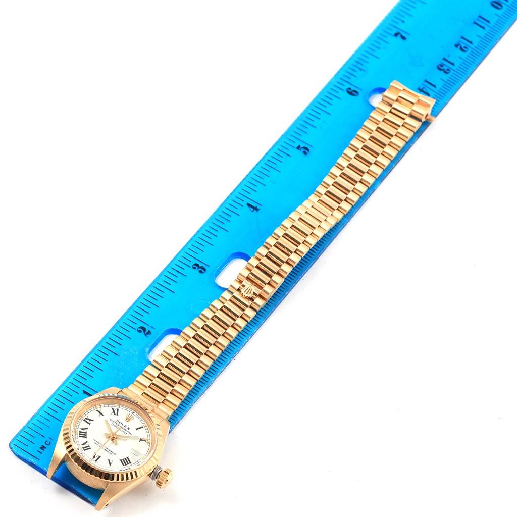 The image shows a top view of a Rolex President 6917 Women's Yellow Gold White Dial 6917 Women's Yellow Gold White Dial model watch placed next to a blue ruler.