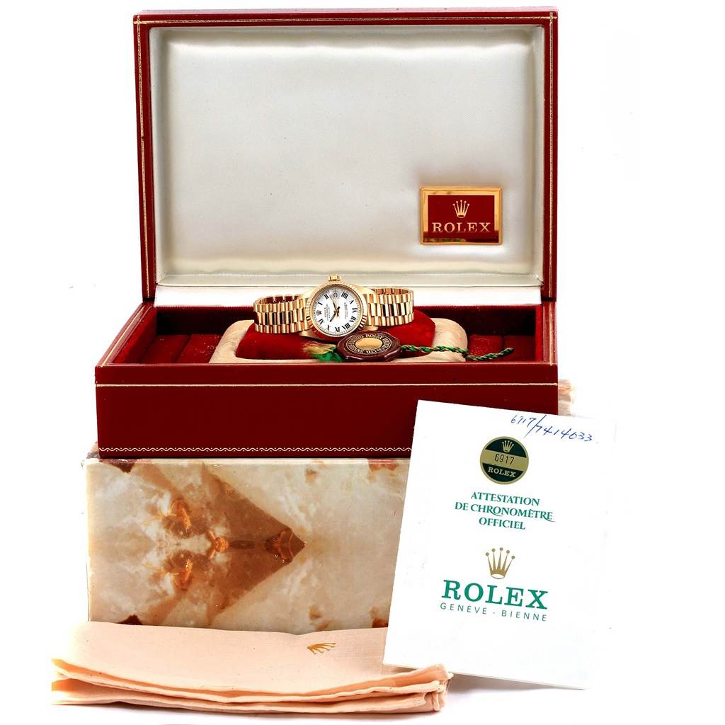 The Rolex President 6917 Women's Yellow Gold White Dial 6917 Women's Yellow Gold White Dial watch is displayed in its box along with a certificate, a tag, and a fabric cloth.
