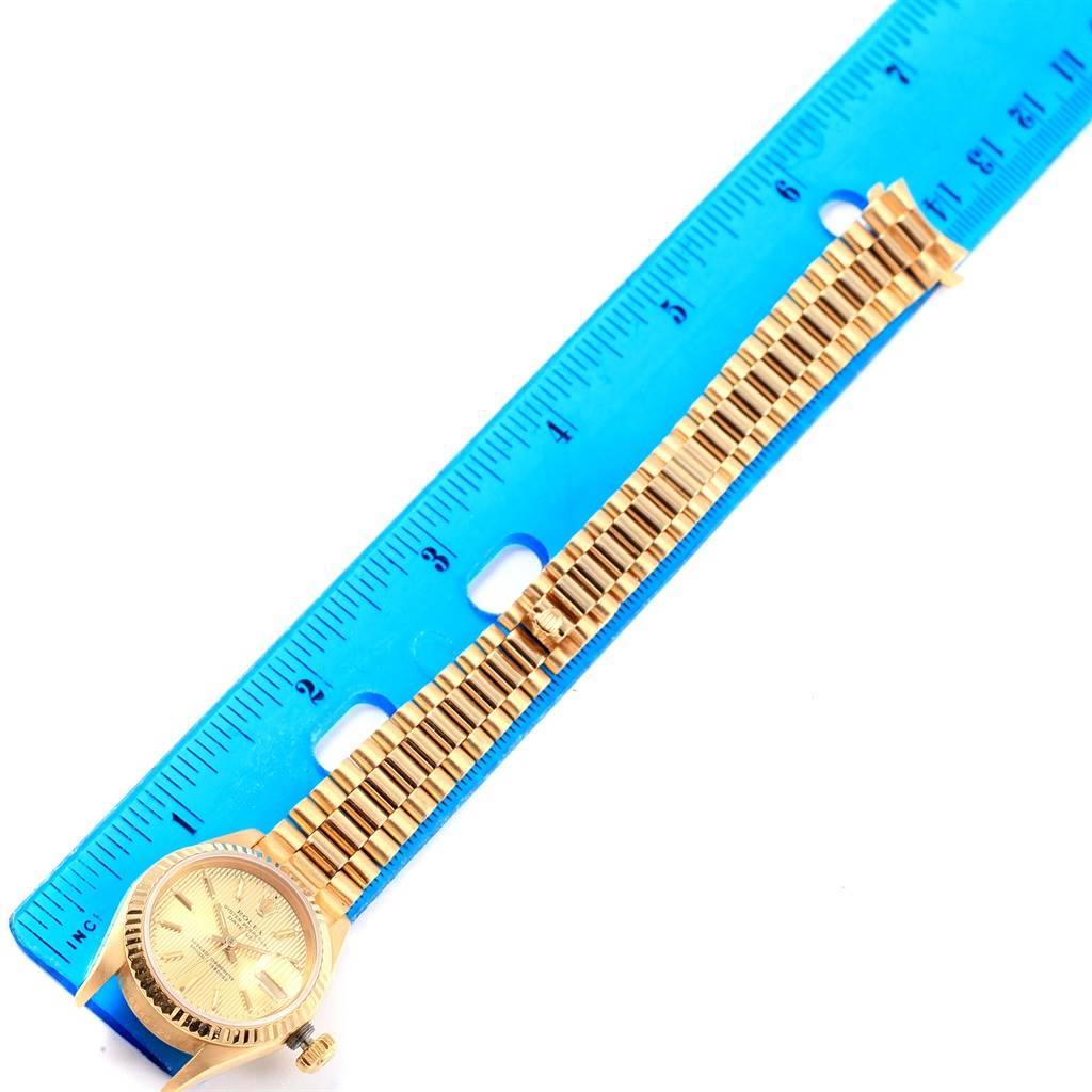 The image shows a Rolex President 69178 Women's Yellow Gold Champagne Dial 69178 Women's Yellow Gold Champagne Dial model watch placed diagonally against a blue ruler, highlighting its length and bracelet links.