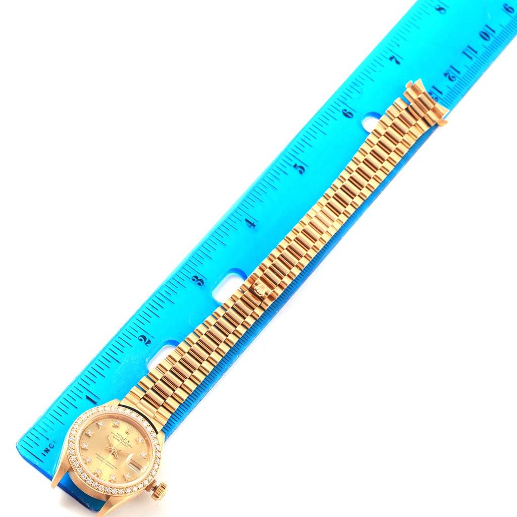 The image shows a gold Rolex President 69138 Women's Yellow Gold Champagne Dial 69138 Women's Yellow Gold Champagne Dial model watch with its band fully extended and placed next to a blue ruler.