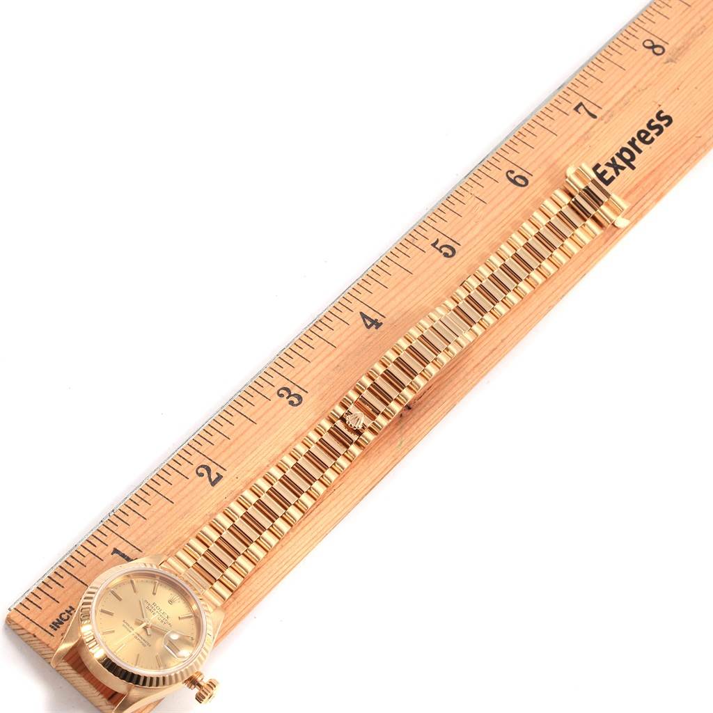 The watch, a President model by Rolex, is shown from a top view, highlighting the face and gold band alongside a ruler for scale.