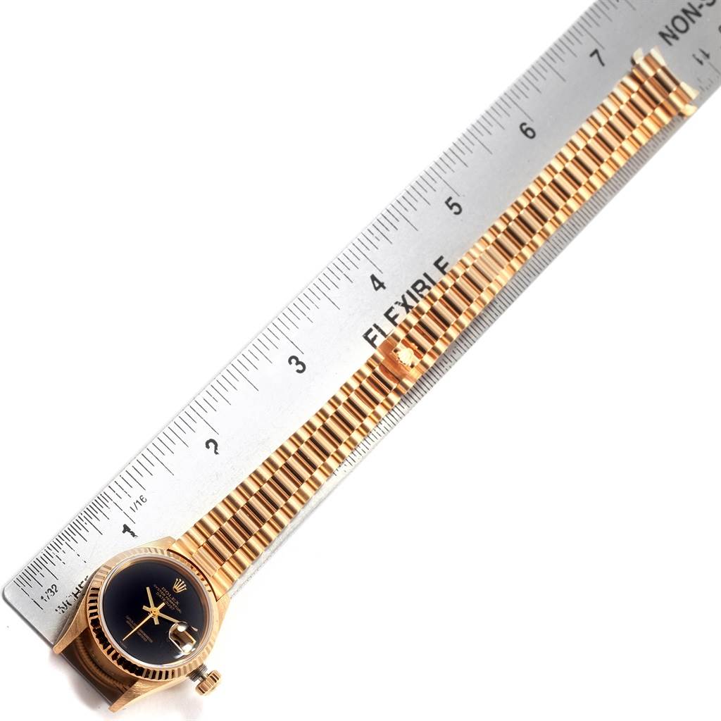 The Rolex President 69178 Women's Yellow Gold Black Dial 69178 Women's Yellow Gold Black Dial model watch is shown face-up next to a ruler, highlighting its gold band and black dial.