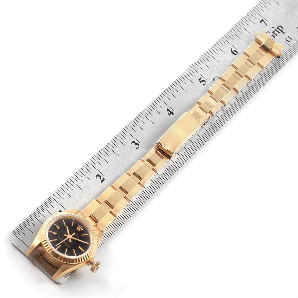 The image shows a Rolex President 67198 Women
s Yellow Gold Black Dial 67198 Women
s Yellow Gold Black Dial model watch lying flat next to a ruler, displaying the watch face and band.