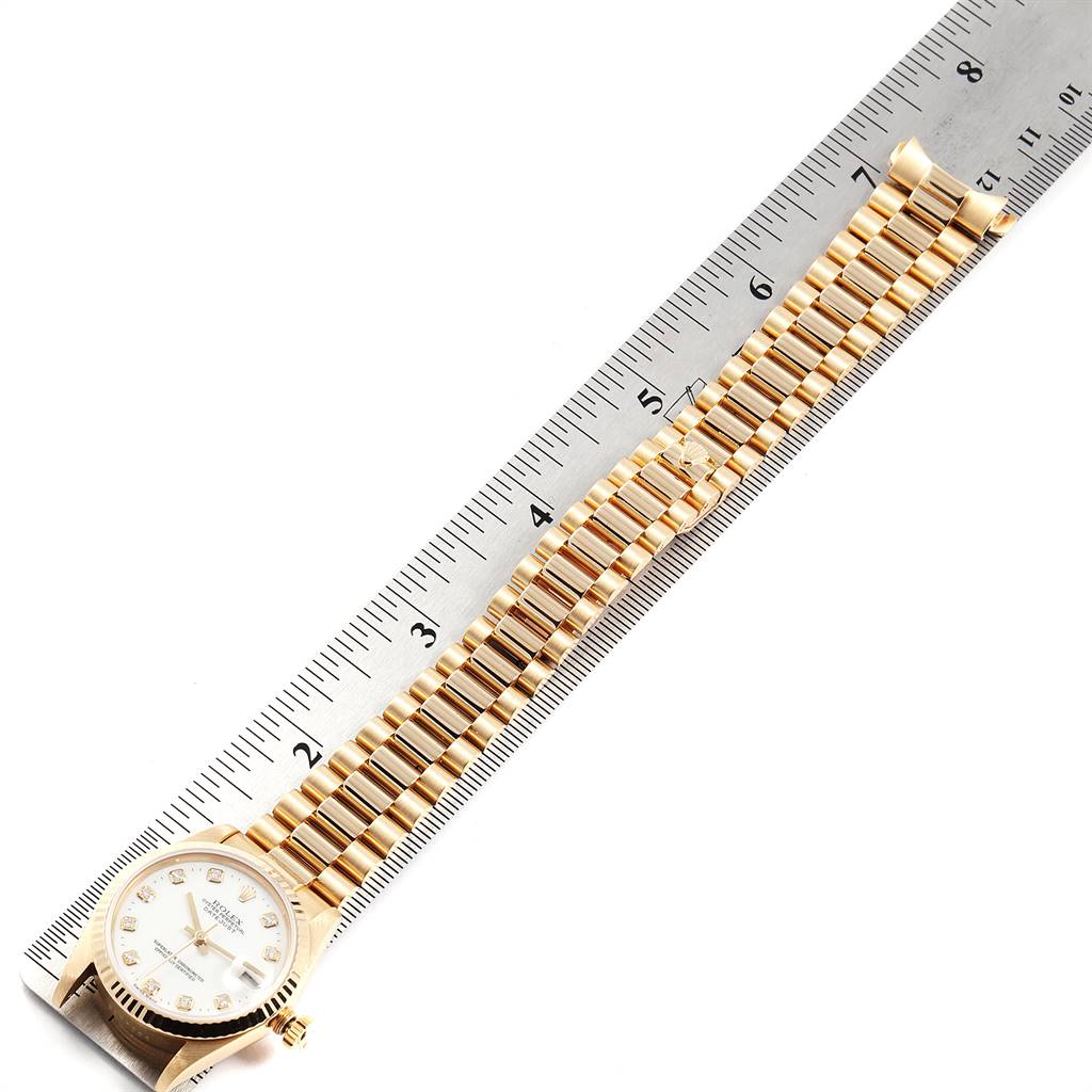 The image shows a Rolex President 68278 Women's Yellow Gold White Dial 68278 Women's Yellow Gold White Dial model watch laid against a ruler, highlighting its gold bracelet and face.