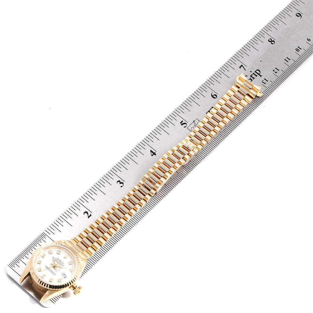 The image shows a Rolex President 69138 Women
s Yellow Gold White Dial 69138 Women
s Yellow Gold White Dial model watch laid flat next to a ruler, showcasing its band and face.