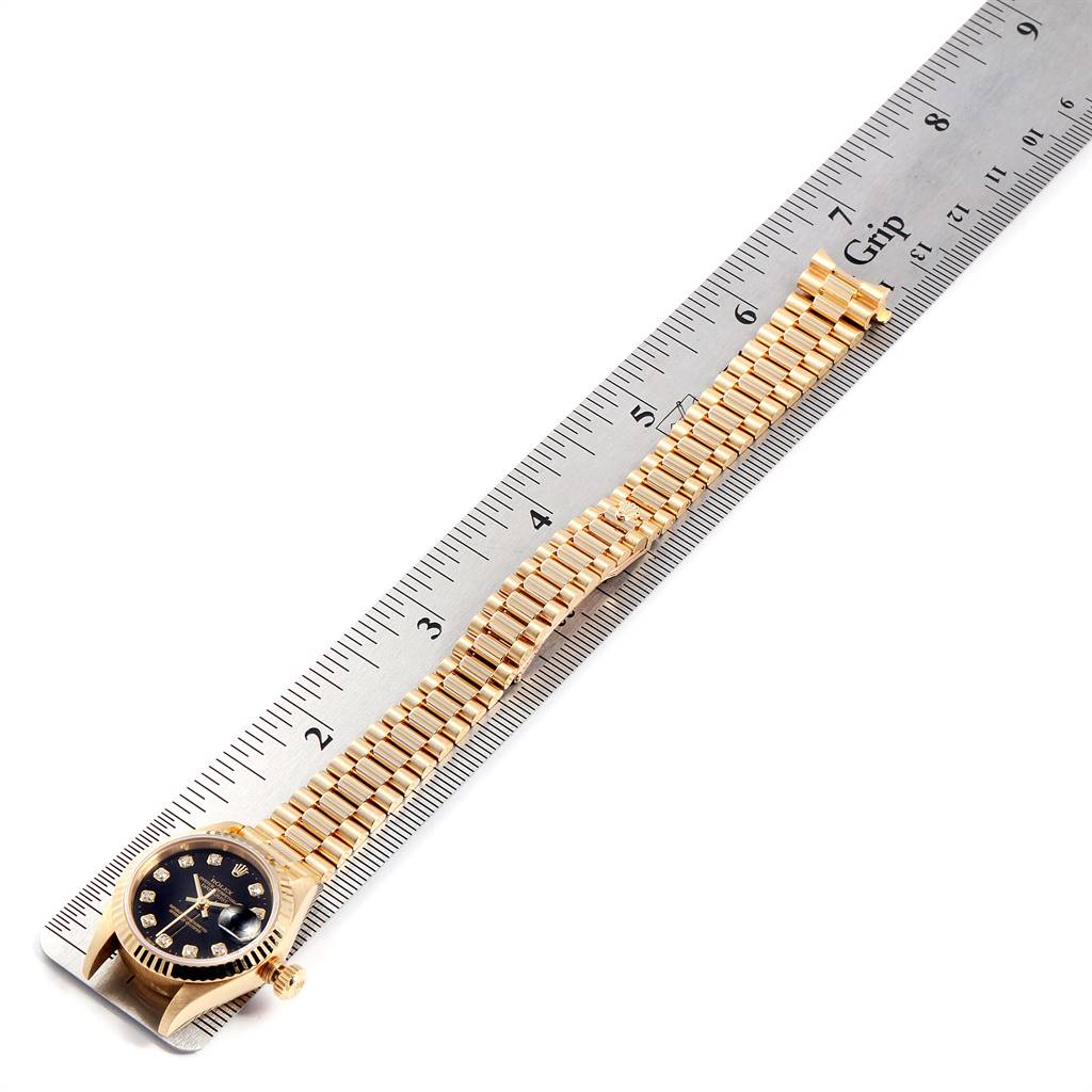 This image shows the Rolex President 79178 Women's Yellow Gold Black Dial 79178 Women's Yellow Gold Black Dial model watch laying flat on a ruler, displaying the case, dial, and gold bracelet.